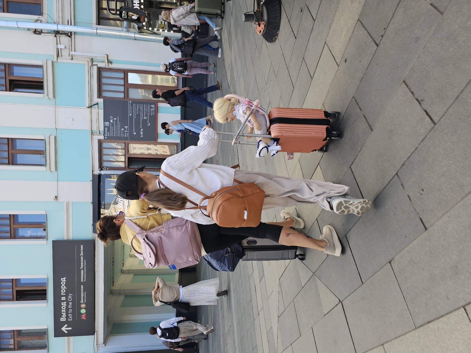 The image depicts a street scene in Moscow, Russia, likely near a metro station or a transit hub, given the signage. In the foreground, two individuals are walking away from the camera. The person on the right is a young girl wearing a white long-sleeved top, light-colored pants, and a small brown backpack. She is pulling a small, coral-colored suitcase with a doll sitting in a stroller attached to the top. The person on the left is an adult woman wearing a yellow jacket, black leggings, and a pink backpack. She is also pulling a large, gray suitcase with wheels.
Several other people are visible in the mid-ground and background, walking in various directions. Some are carrying backpacks or luggage. The architecture suggests an urban environment with light blue and white building facades and large windows. A sign with Russian text, including "Выход в город" (Exit to the City) and "Exit to the City" in English, is prominently displayed on the building. Other signs in Russian indicate metro lines and directions, such as "Белорусская" (Belorusskaya) and "Площадь Тверская Застава" (Tverskaya Zastava Square). The ground is paved with large, gray stone tiles. A street cleaning machine with brushes is visible on the right side of the image. The overall atmosphere is that of a busy day in a public urban space.