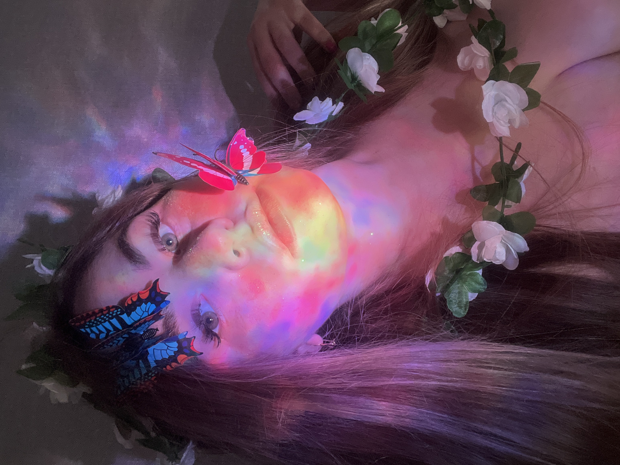 A person is lying down with their face directed toward the viewer. They have long brown hair adorned with a garland of white flowers and green leaves. Two artificial butterflies are attached to their face; a red one on their forehead and a blue one near their eye. The person is illuminated by a multicolored light, casting hues of pink, orange, and blue across their face. Their eyes are open, and their skin appears to have glitter on it. The background is dark and indistinct. The photograph appears staged. The location, Chaikovsky, Russia, is not determinable from the image.