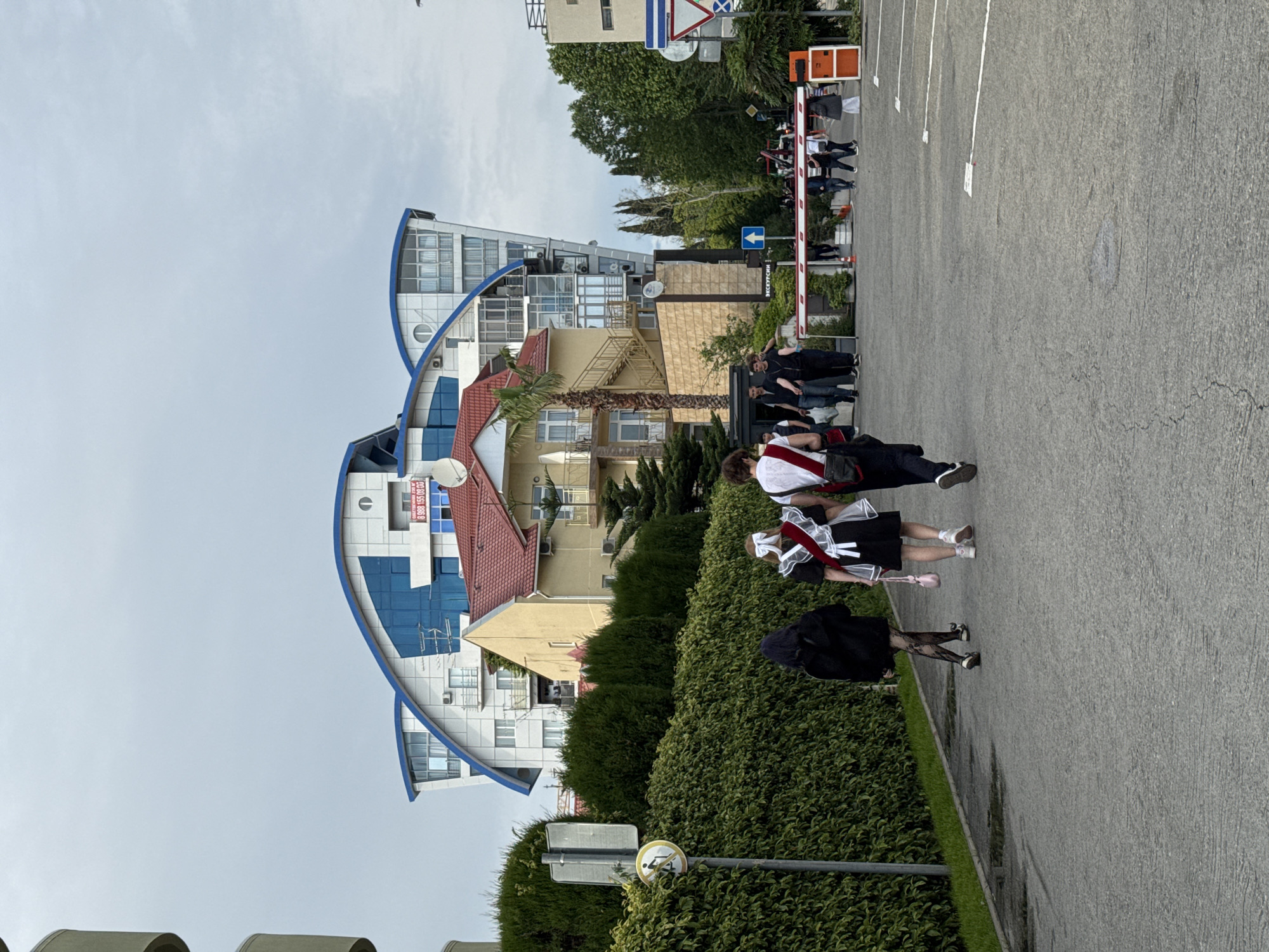 The image depicts a street scene in Sochi, Russia. In the foreground, three people are walking on a sidewalk beside a parking lot. Two of them appear to be wearing school uniforms with white aprons and bows. In the background are several buildings, including a modern building with blue accents. A crowd of people is visible behind a barrier in the distance. The sky is overcast.
