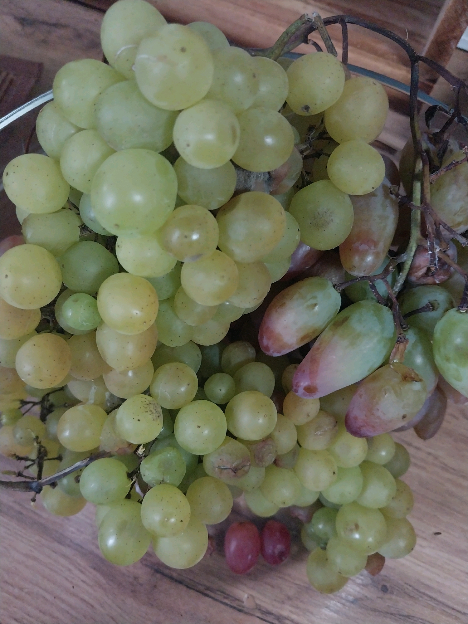 This is a close-up, overhead shot of a bunch of green and some reddish-purple grapes resting on a wooden surface. The grapes are clustered together, with some vines visible. The wooden surface appears to be a tabletop or counter, with visible grain patterns. There are no people or other objects in the frame. The lighting is natural and appears to be from above, casting soft shadows. The scene is not specific to any location in Dnipro, Ukraine; it is a generic still life image of grapes.
