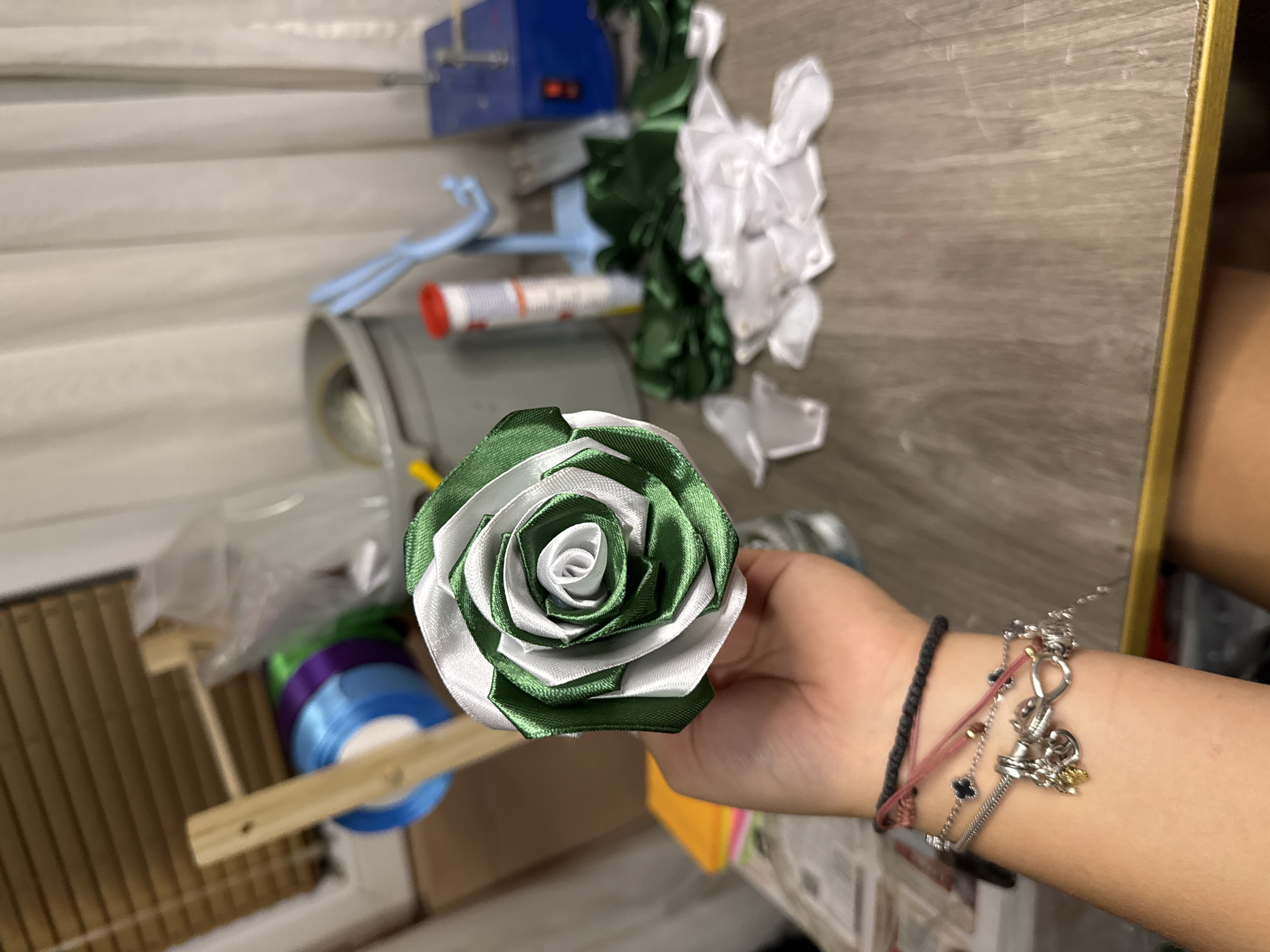 A hand holds a green and white ribbon rose, its petals tightly wound towards the center. The background is a workshop or craft room, with spools of ribbon, fabric scraps, and various tools visible. The scene appears to be indoors, with a wooden table in the foreground and blinds on a window in the background. There are no discernible landmarks or architectural features that could identify the location as Sredets, Bulgaria.