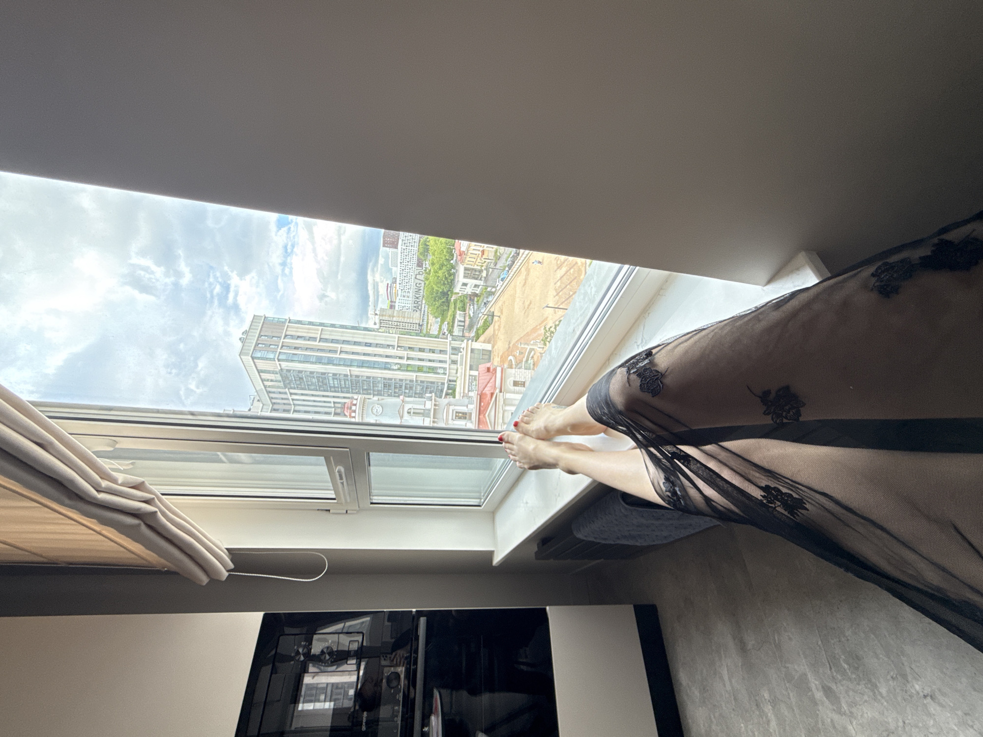A low-angle shot depicts a person with painted red toenails resting their feet on a windowsill. The person is partially covered by a black, sheer, floral-embroidered fabric. Outside the window is a view of buildings and streets in Dolgoprudny, Russia. In the foreground are parts of the room's interior, including closed light-colored blinds, a black and white appliance, and a gray wall.