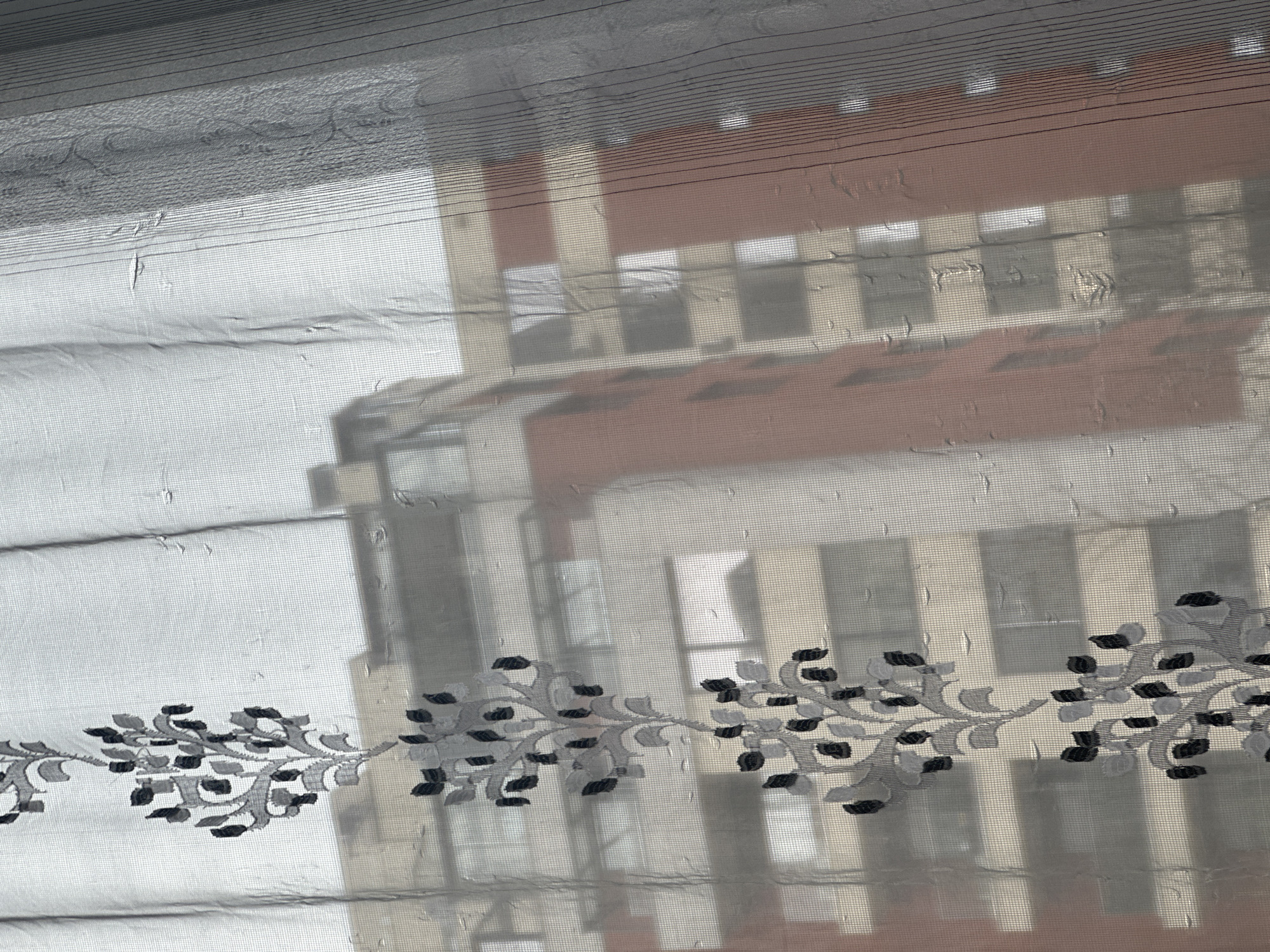 A close-up shot through a sheer curtain with a floral design captures a blurry view of a building. The building appears to have multiple stories with a mix of white and light brown exterior walls. Windows are visible on each level. The curtain has a horizontal striped pattern at the top. The floral design on the curtain consists of branches with leaves in gray and black hues. The exact location in Varna, Bulgaria, cannot be determined from the image.