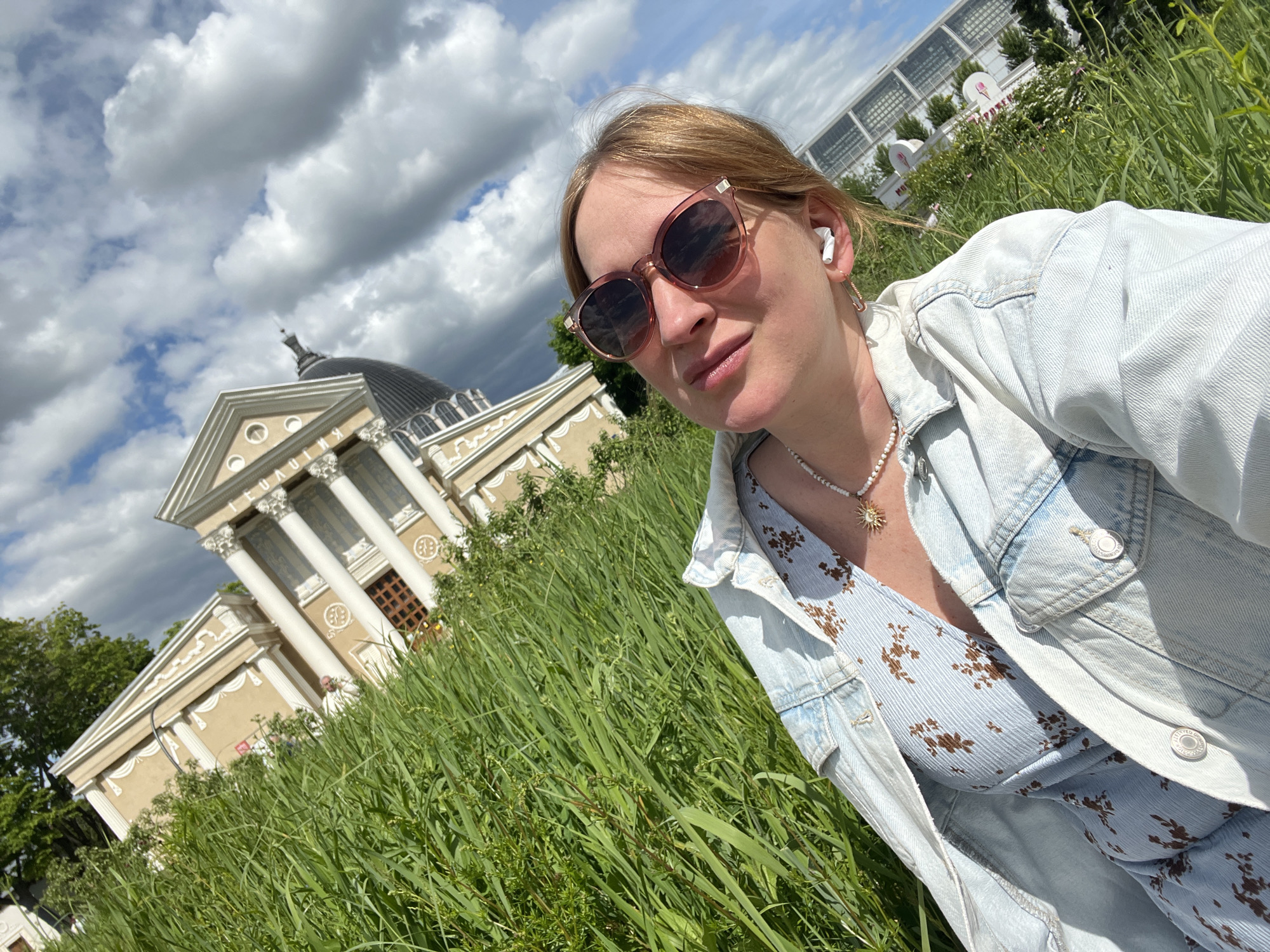 A fair-skinned woman wearing sunglasses, a denim jacket, and a light blue floral dress takes a selfie in a field of tall green grass. She has blonde hair pulled back, wears a white earpiece, and a necklace with a sun pendant. Behind her to the left is a pale tan building with white columns and a dark dome. In the distance on the right is a building with a white roof and glass walls. The sky is partly cloudy with bright light. The photo was taken at the All-Russia Exhibition Center in Moscow, Russia.