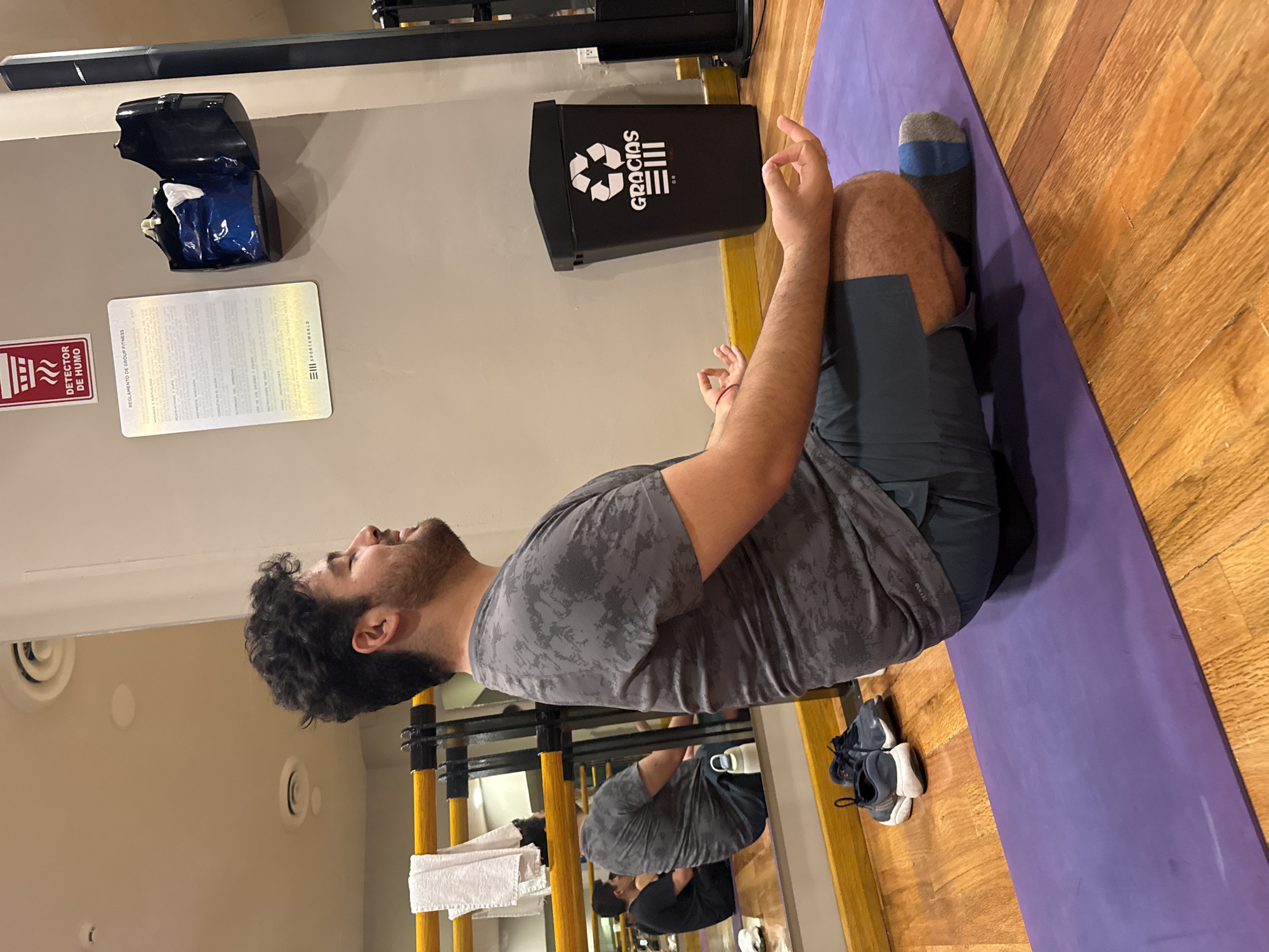 A man with dark curly hair and a beard sits cross-legged on a purple yoga mat on a wooden floor. He wears a gray patterned t-shirt and dark gray shorts, with gray and blue socks visible on his right foot. His hands are raised in a meditative gesture. Behind him is a gray wall with a black recycling bin labeled "GRACIAS" and a fire safety sign. Above the bin is a blue and black object. A sign is also posted on the wall. To the left of the man is a mirror reflecting his image and a yellow structure with towels. A pair of athletic shoes sits beside the yoga mat. The location is a fitness studio or gym, potentially in Puebla, Mexico, considering the "GRACIAS" label and signs in Spanish.