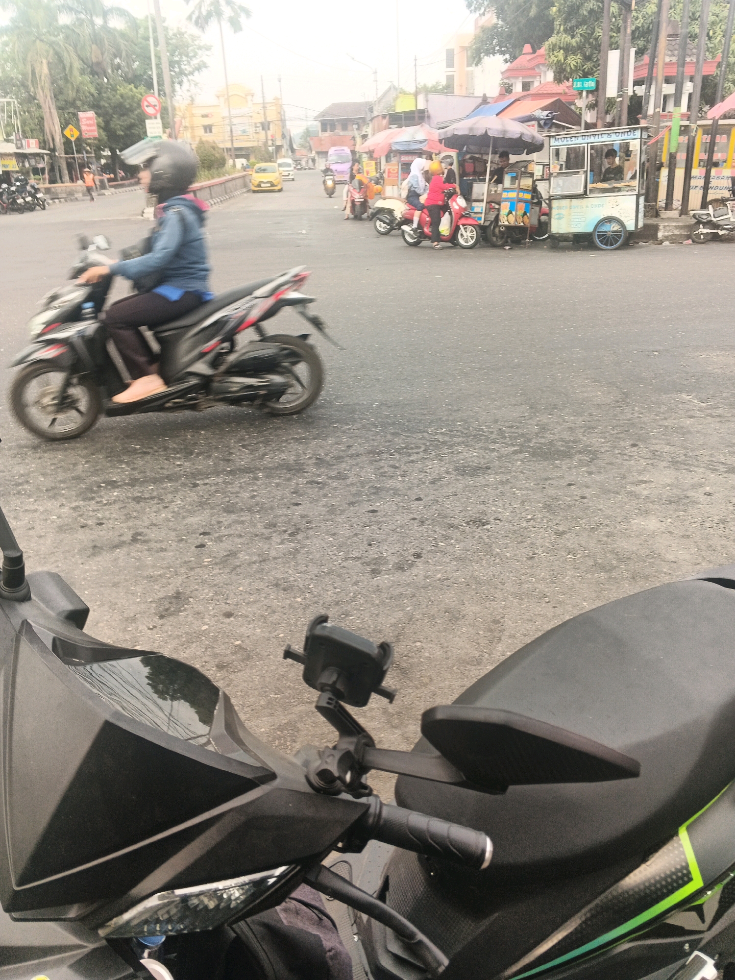 A low-angle, slightly blurry, eye-level shot captures a street scene in Ungaran, Indonesia. The foreground is dominated by the front portion of a dark-colored scooter, its handlebars and a phone mount visible. In the midground, a person wearing a blue jacket and helmet rides a black and red scooter, their right leg extended. The scooter is in motion. Behind them, a red scooter carries two passengers, one in a red outfit. To the right, two food carts are parked alongside the road. One cart, a light blue and white color, has the words "MOLEK UNYIL & ONDE" written on it. The other cart, partially obscured, is covered by a dark umbrella. Various buildings with red roofs are visible in the background, along with palm trees and utility poles. The road surface is asphalt, appearing worn and patched. A yellow car is visible further down the street. The overall impression is that of a busy, everyday street scene.