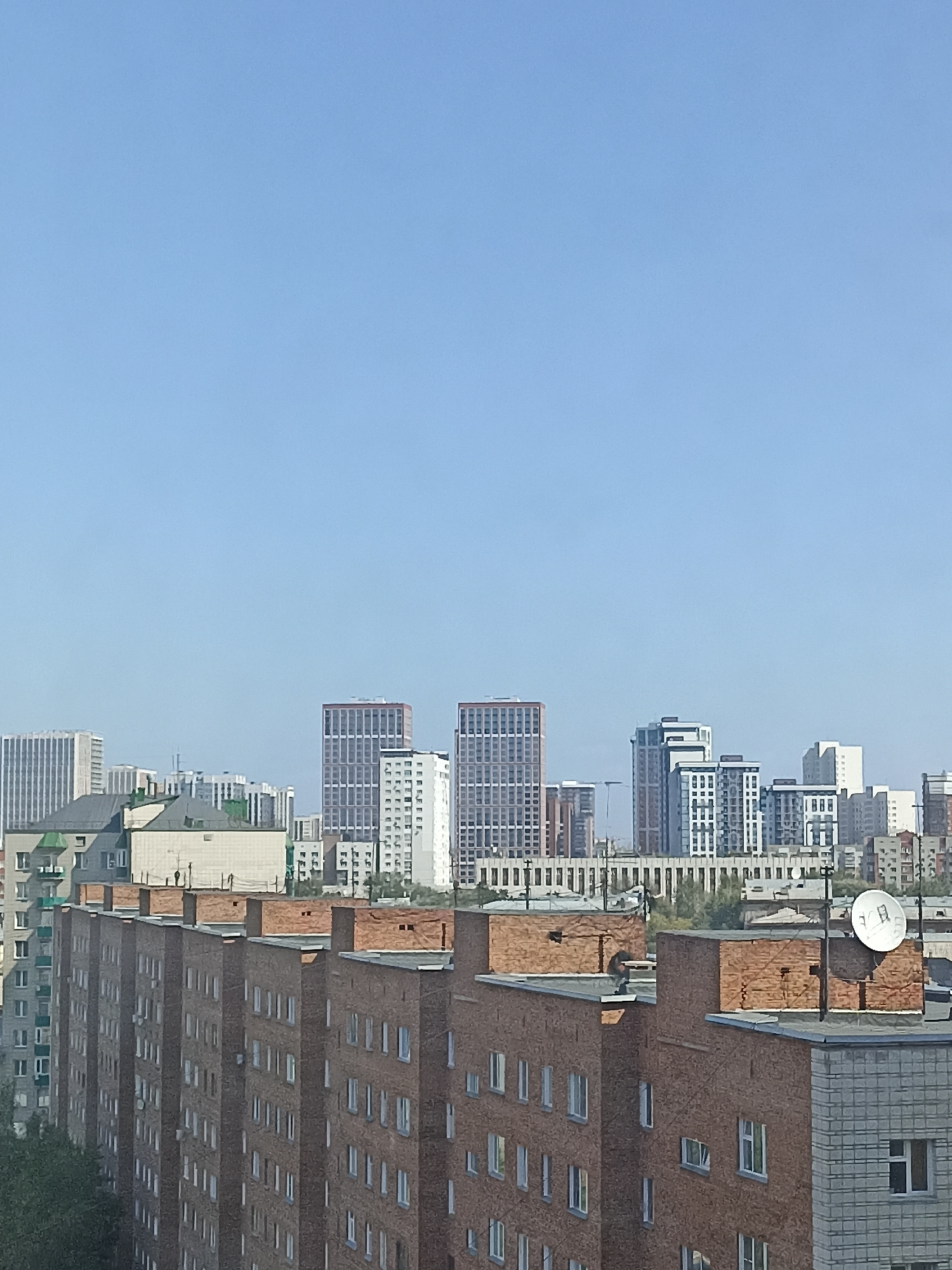 The image shows a cityscape in Novosibirsk, Russia, under a clear blue sky. In the foreground, there are several brick apartment buildings with visible windows and chimneys. Behind these, a line of more modern, taller buildings, including some with distinctive patterned facades, forms the city skyline. Further in the background, more residential and office buildings can be seen. A satellite dish is mounted on the roof of one of the brick buildings. The overall impression is of a residential and business district.