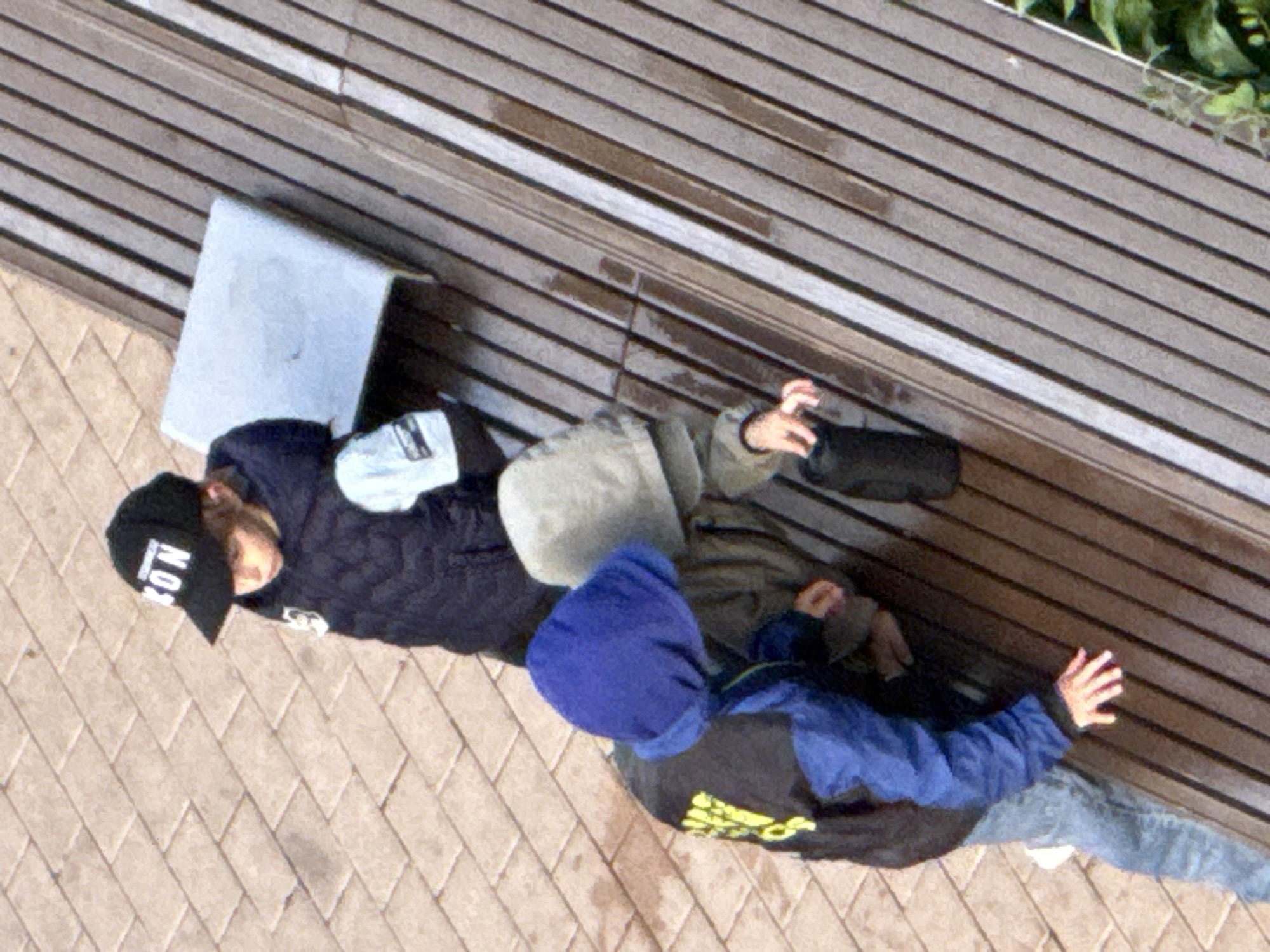 Three children are positioned on a wooden slatted bench and the adjacent paved area. The child on the left is wearing a dark blue puffer jacket with a black hood, blue sleeves, and blue jeans. They have a blue beanie on their head and their left hand is resting on the bench. The child in the middle is wearing a light gray hooded jacket. The child on the right is wearing a dark blue puffer jacket with a black cap that has white lettering. A light blue rectangular object is on the bench. The ground is paved with light brown rectangular tiles. The overall scene appears to be outdoors, possibly in a park or public area in Moscow, Russia.