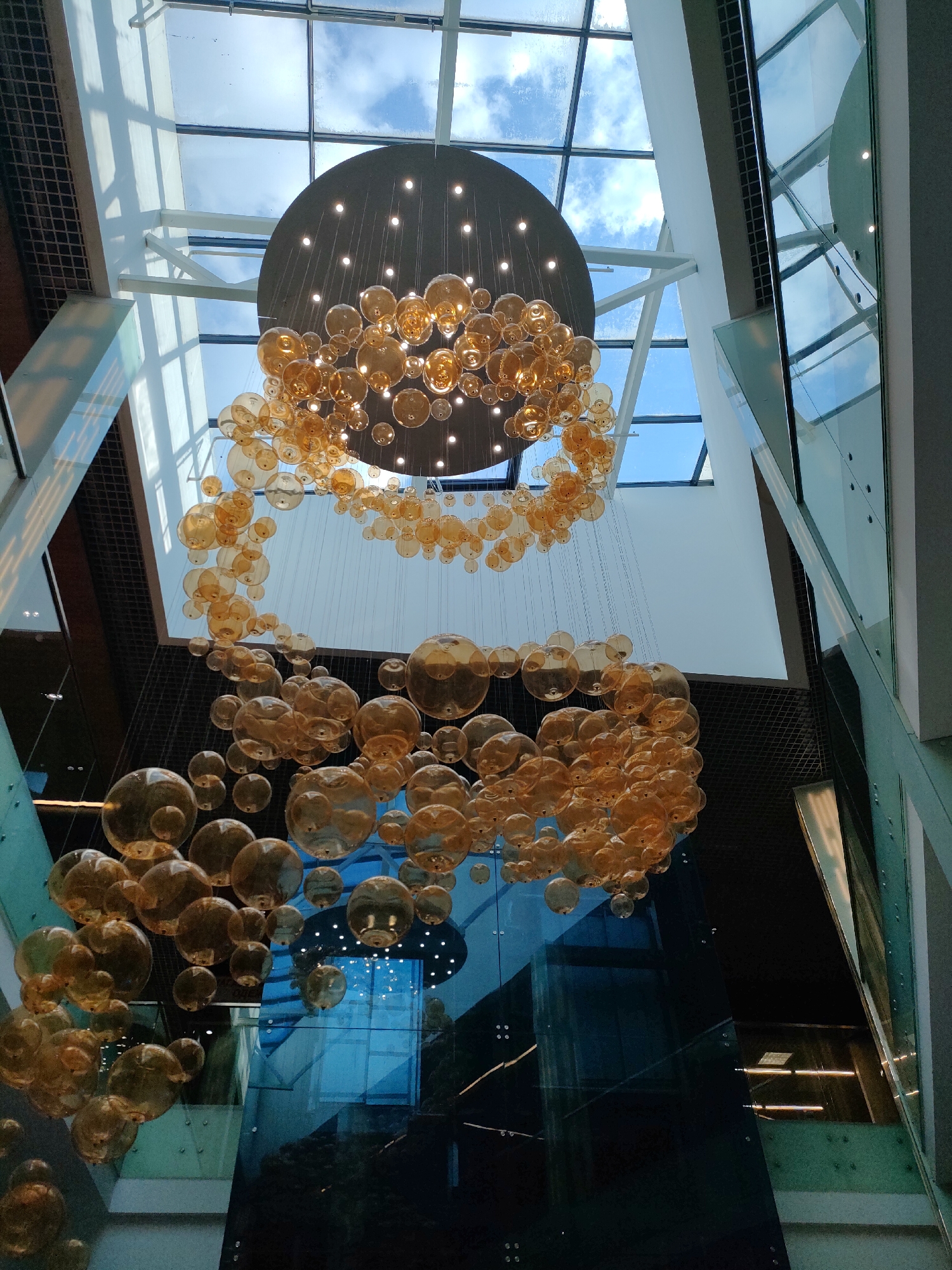 The image shows a large, modern atrium with a dramatic chandelier. The chandelier is composed of numerous amber-colored glass spheres of varying sizes, suspended from a circular black fixture. The spheres are clustered and cascade downwards, creating a dynamic and eye-catching focal point. Above the chandelier, a large glass ceiling allows natural light to filter in, revealing blue sky and clouds. The atrium features sleek architectural elements, including glass walls and metallic structural beams, suggesting a contemporary commercial or public space.
This scene is located in the "Dana Mall" in Minsk, Belarus. Specifically, it is the central atrium of the mall, characterized by its multi-story design and the prominent hanging art installation. The lighting in the image suggests it was taken during daylight hours. There are no people visible in this particular frame. The environment is clean and well-maintained, consistent with a shopping mall setting.