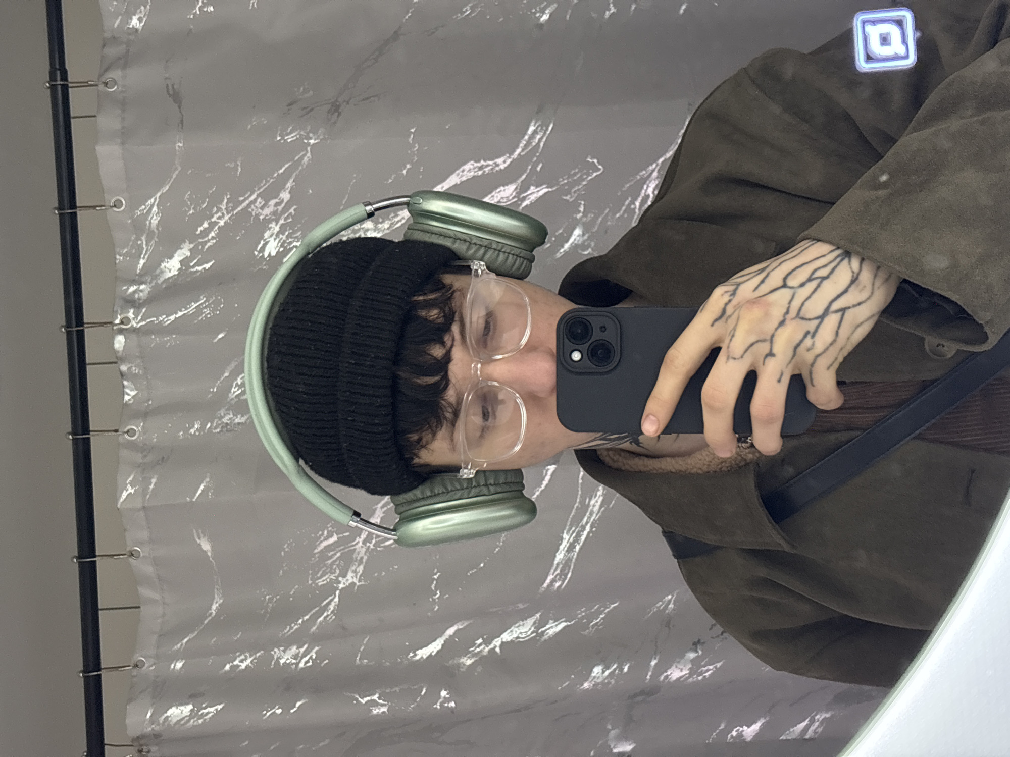 The image displays a mirror selfie taken indoors within the city of Saratov, Russia, specifically in what appears to be a bathroom. The subject, an individual, is reflected from the chest up, wearing a black knit beanie and clear-framed eyeglasses. Mint green over-ear headphones are worn over the beanie. The individual wears a dark olive green or brown textured jacket with a prominent collar, revealing a brown inner layer and a light-colored, possibly shearling-like, lining around the neck area. The left hand, adorned with extensive black line-work tattoos resembling veins or branches and smaller finger tattoos, holds a black smartphone (likely an iPhone) with its camera lenses pointed towards the mirror to capture the reflection. A minor reddish mark is present on the back of the left hand. The background consists of a crinkled, translucent white shower curtain suspended from a dark rod by silver hooks. An illuminated blue square icon with a stylized symbol is visible on the right side, suggesting a modern fixture or control.