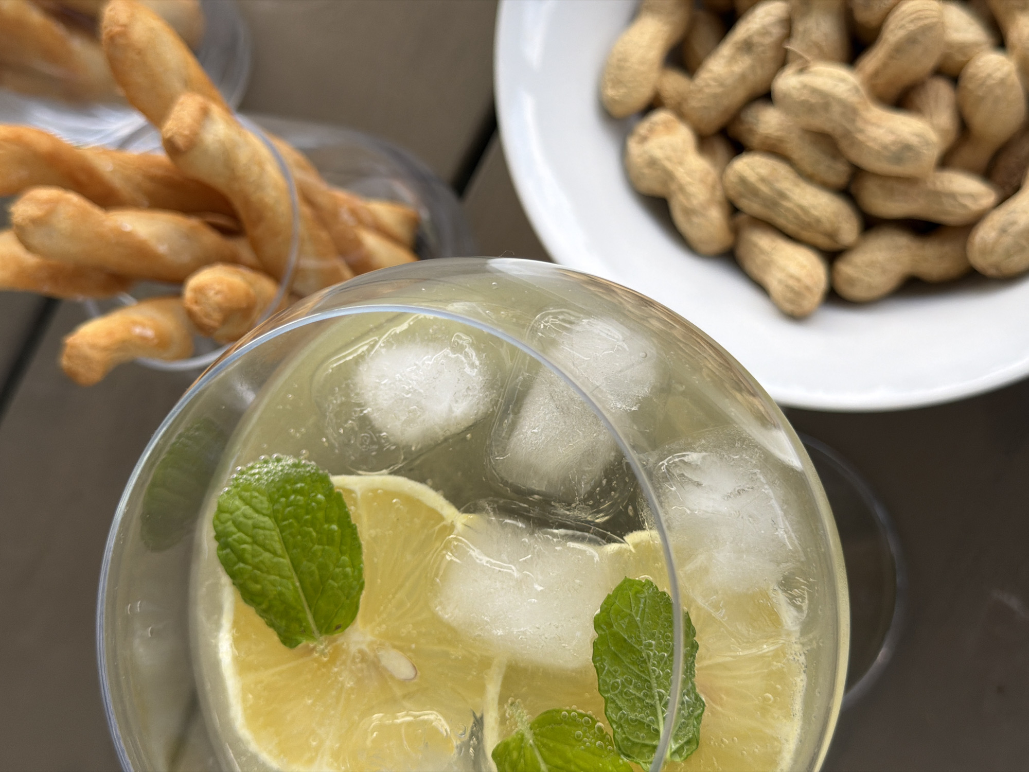 A close-up, overhead shot shows a refreshing drink in a wide-rimmed glass. The drink is clear with a slight yellow tint, filled with ice cubes, lemon slices, and garnished with fresh mint leaves. To the right of the drink, a clear glass is filled with golden-brown breadsticks. In the bottom right corner, a white bowl overflows with unshelled peanuts. The objects are placed on a dark, textured surface, possibly a table. The scene appears to be set outdoors, given the lighting and natural elements. No people are visible. The context of this scene in Junglinster, Luxembourg, is not determinable from the image alone.