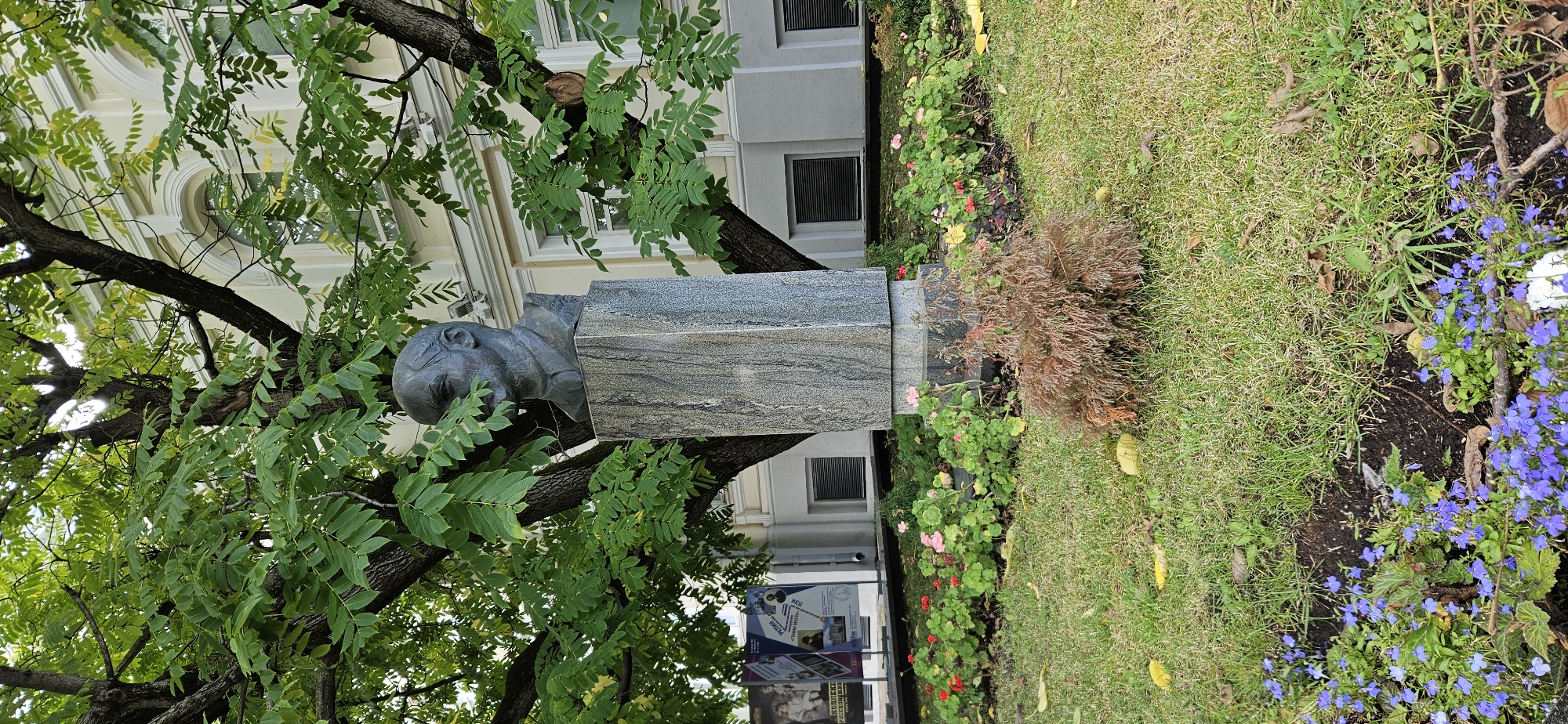 The photograph depicts a bust of a man, likely a historical figure, positioned on a grey marble pedestal. The bust and pedestal are situated outdoors, beneath the canopy of a large tree with abundant green leaves. The background reveals a building with arched windows, suggesting an urban setting. In the foreground, a bed of small red and pink flowers is visible, along with patches of green grass and fallen yellow leaves. Further down, a cluster of small blue flowers is blooming. The overall impression is of a monument in a park or garden area within a city. Based on the context provided, this scene is identified as being located in the city of Vitsebsk, Belarus.