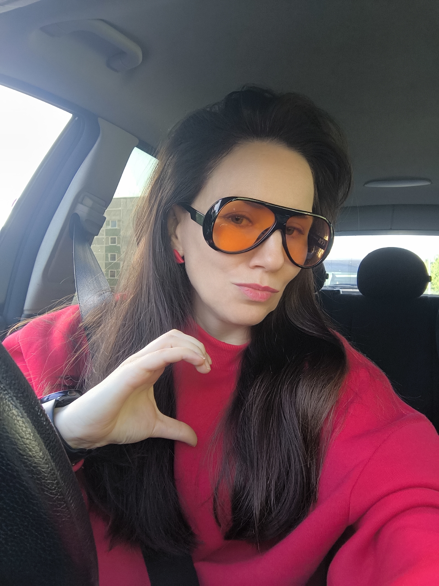 A woman is inside a car wearing orange aviator sunglasses and a red sweatshirt. She has long, dark hair and red earrings. She is making a heart gesture with her hand. The car's interior is visible, with a black steering wheel and seats. Through the car window, there is a building with a light-colored facade, potentially in Riga, Latvia. The background suggests a city environment during daylight.