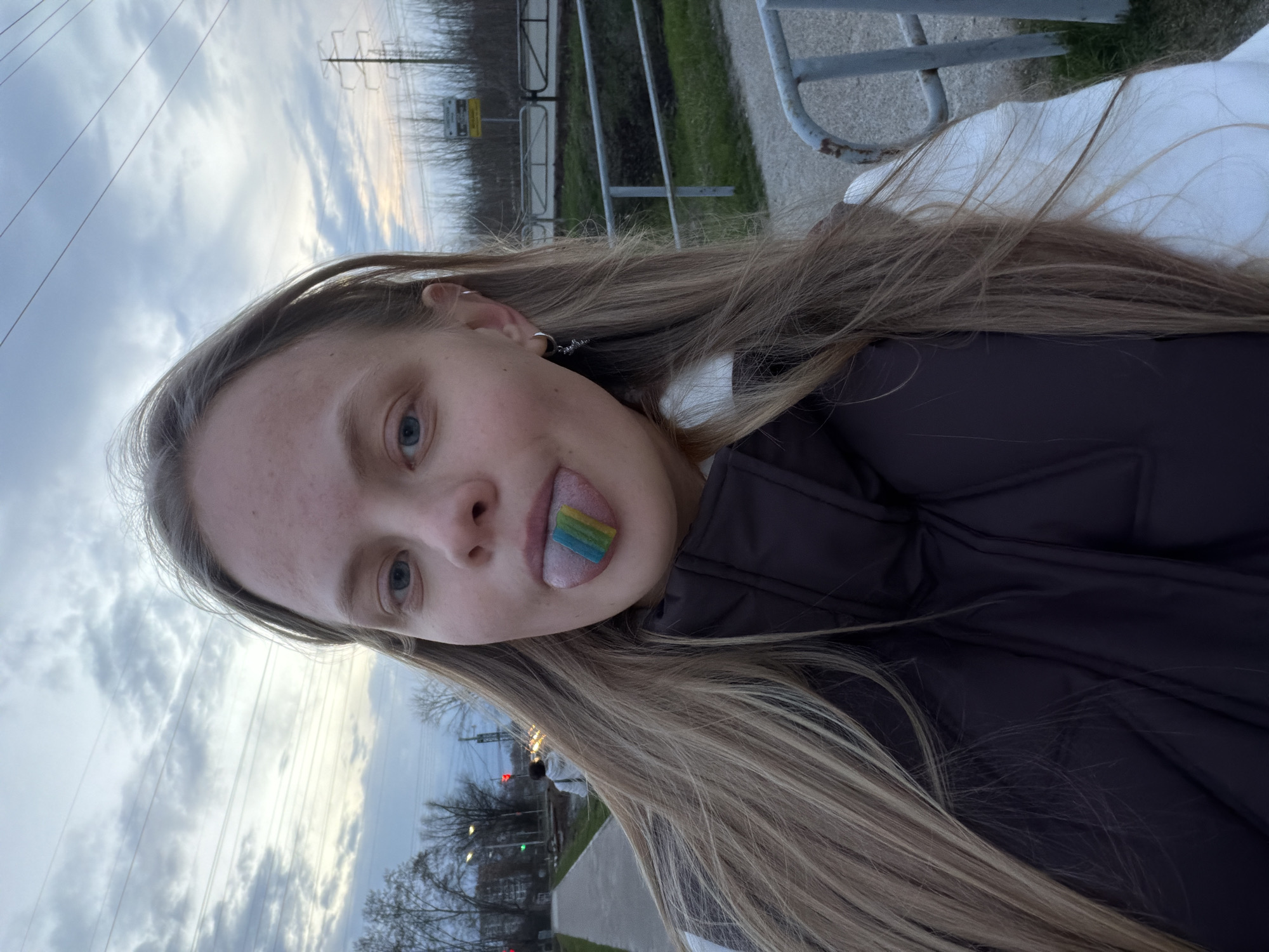 A young individual, likely female, is featured in a close-up, slightly tilted portrait, taken outdoors in what appears to be Chaikovsky, Russia. The subject has long, light brown hair, light blue eyes, and freckles visible on their forehead. They are wearing a dark, possibly black or navy, puffy jacket over a white layer, and a small silver hoop earring with a dangle is visible in their left ear.
The central activity captured is the person playfully sticking out their tongue, on which rests a small, rectangular, multicolored candy. The candy has distinct layers of blue, green, yellow, and orange/red.
The background shows an overcast sky with patches of brighter light, suggesting either late afternoon or early evening, or a generally cloudy day. Visible environmental details include prominent power lines stretching across the frame, utility poles, and a grey metal railing or fence. A concrete path or road is discernible, alongside patches of grass and distant trees with bare branches, indicating a cooler season. In the far background, blurred elements suggest urban or suburban structures. A small sign on one of the utility poles has light-colored, unreadable text on a yellow or white background. The overall mood is casual and playful, highlighted by the person's expression.