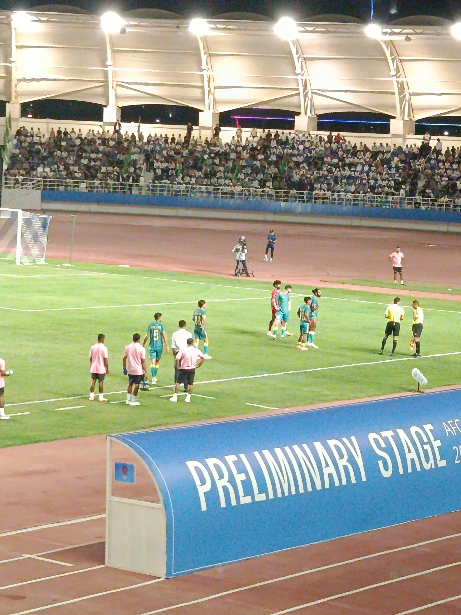 The image shows a nighttime outdoor soccer match at the Arkadag Stadium in Arkadag, Turkmenistan. Spectators fill the stands under stadium lights. On the field, several players in green and pink uniforms are visible, along with two referees in yellow and black uniforms. A soccer goal is set up on the left side of the field. In the foreground, a blue dugout with "PRELIMINARY STAGE" written on it is visible. The stadium features a covered grandstand with white architectural elements.