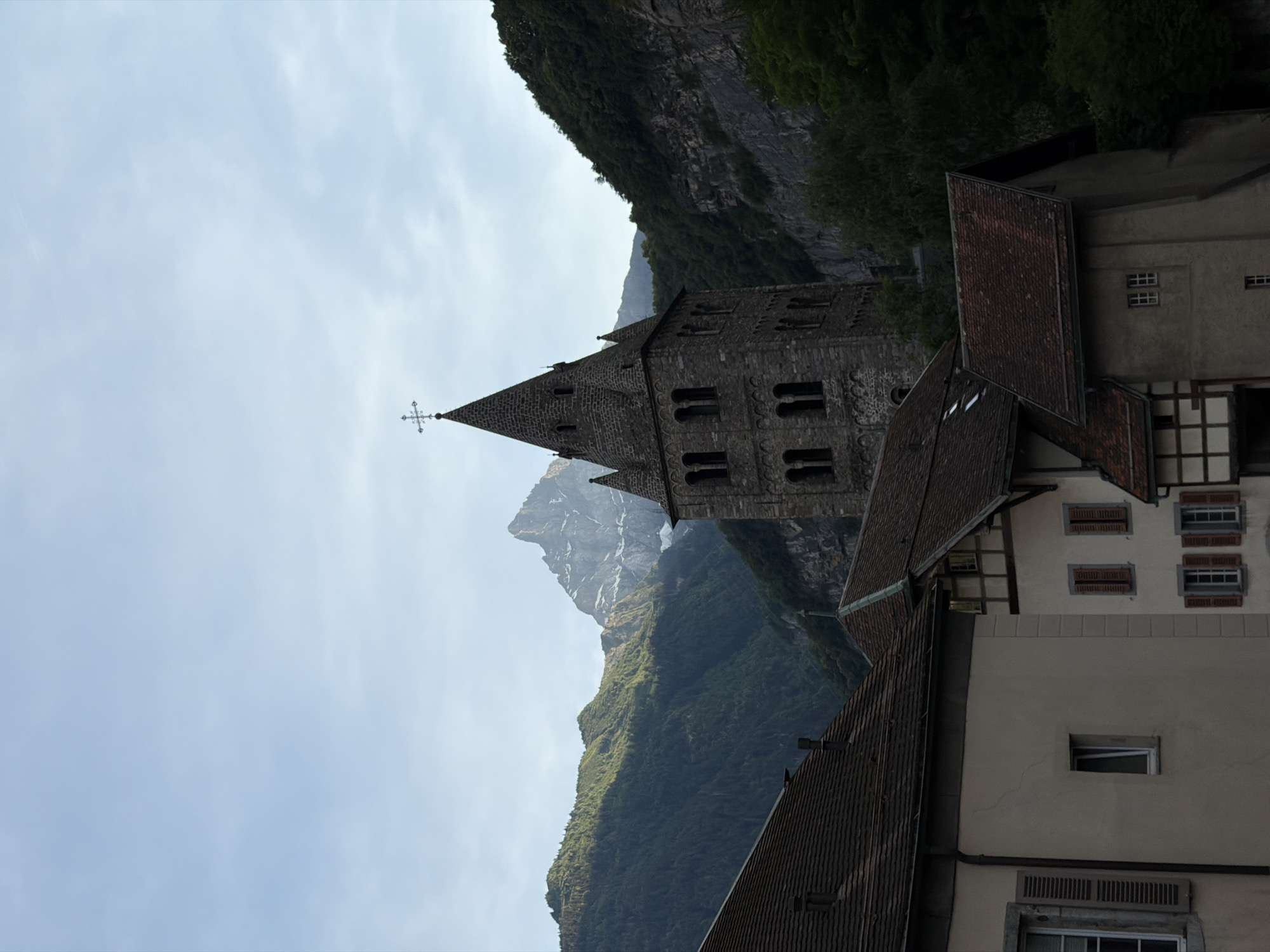 The image shows a view of the Saint-Maurice Abbey in Switzerland. The abbey, built with grey stone, features a tall, pointed spire topped with a cross. The architecture suggests a historical religious site. In the foreground, various buildings with tiled roofs are visible, some with arched windows and shutters, contributing to the rustic charm of the scene. Behind the abbey, a steep, green mountainside rises sharply, partially shrouded by trees. A rocky peak, possibly snow-capped, emerges in the distance, hinting at the alpine environment. The sky is overcast with a pale, diffused light, suggesting it is either early morning or late afternoon, or a generally cloudy day. The overall impression is one of tranquility and historical significance, set against a dramatic natural landscape. There is no visible text in the image.