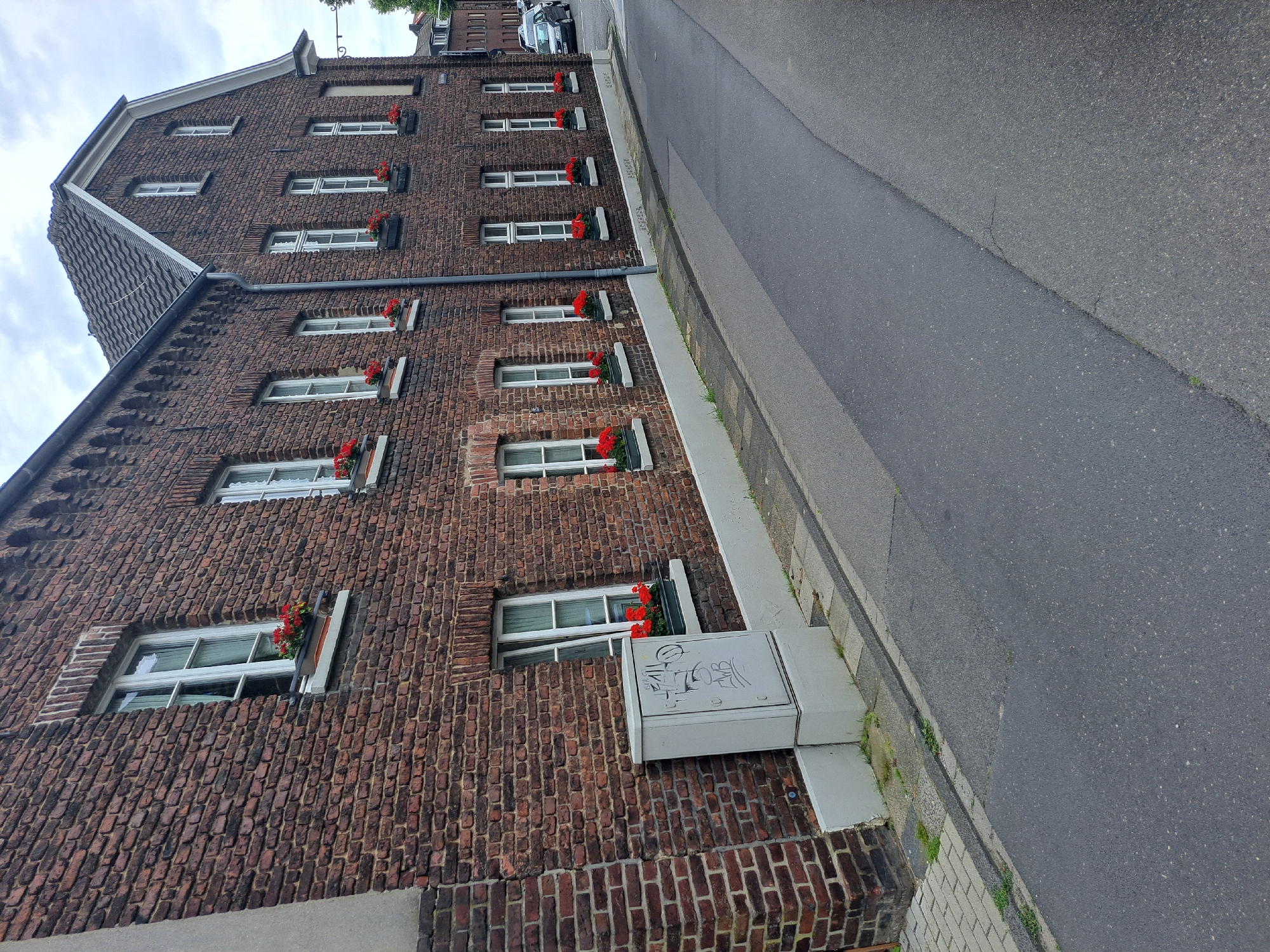 An outdoor, eye-level, medium shot captures a brick building with several windows, each adorned with red flowers in window boxes. A grey utility box, covered in graffiti, is situated at the base of the building. The building is adjacent to an asphalt road with a narrow sidewalk. The sky is overcast. The scene is devoid of people.