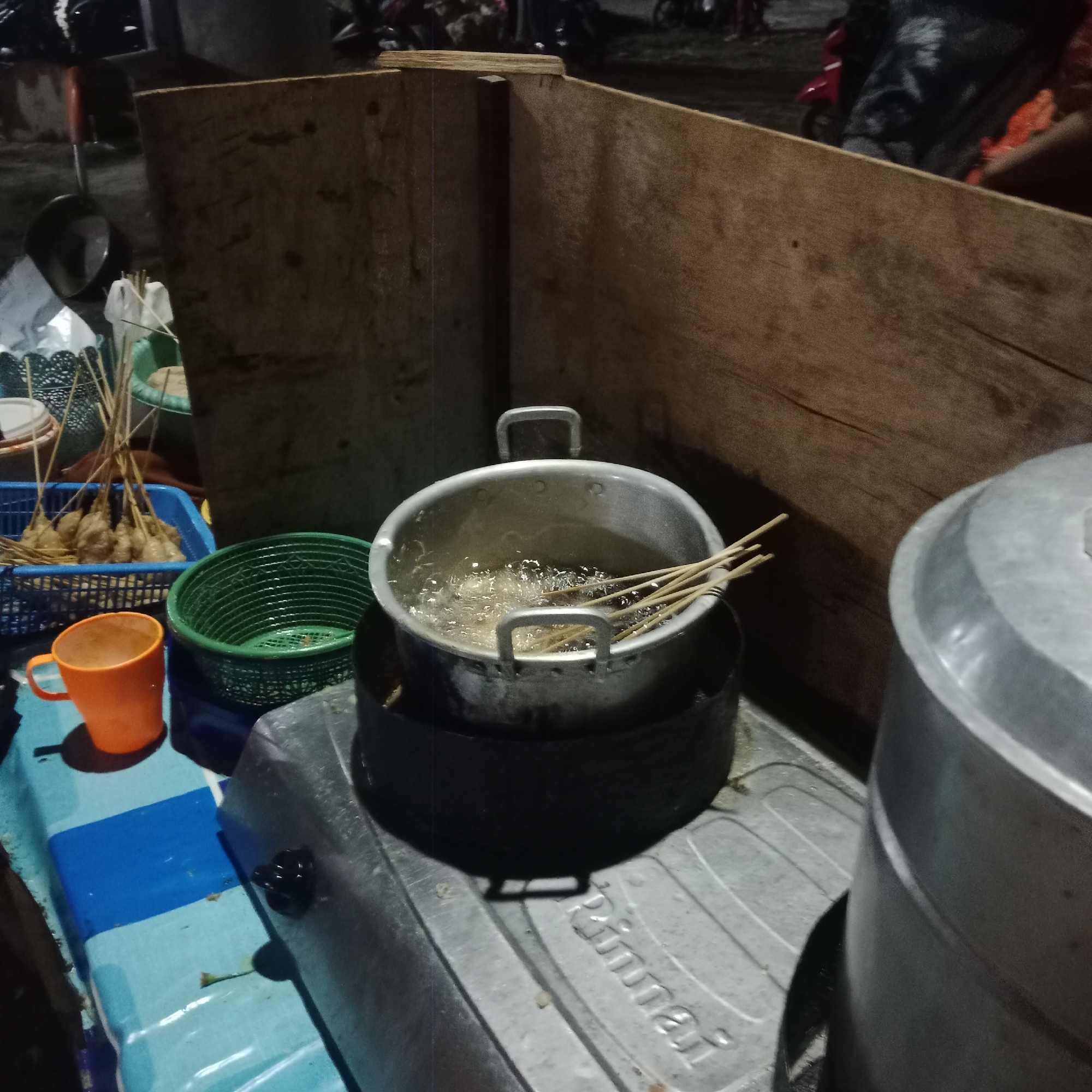 The image depicts an outdoor food stall in Kunjeng, Indonesia, at night. The foreground features a portable gas stove with a metal pot actively frying food. Skewers of food, possibly meatballs or fried dough, are submerged in the bubbling oil. To the left, an orange plastic cup sits on a blue and white striped surface, next to a green basket containing more food skewers. In the background, a wooden partition separates the stall from the dark, outdoor environment where motorcycles and indistinct figures are visible, suggesting a busy street or market. The overall impression is that of a humble, functional food preparation setting operating in the evening.