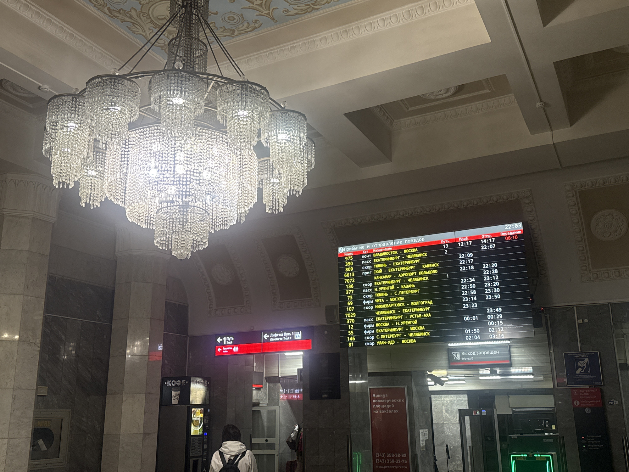 The image depicts the interior of the Yekaterinburg railway station in Russia. A large, ornate crystal chandelier dominates the upper part of the frame. Below, a departure board shows train schedules. The board displays the train number, category, destination, platform number, arrival time, departure time, and delay information for various trains. To the left, there is a coffee vending machine and a sign indicating directions to "track 1", including the location of the lavatory. A person with a backpack is visible in the foreground, seemingly waiting or passing through the station. Further back, another individual is standing near what appears to be a security screening area. Commercial space lease advertisement visible. The environment suggests a public transportation hub with elements of classic architectural design.