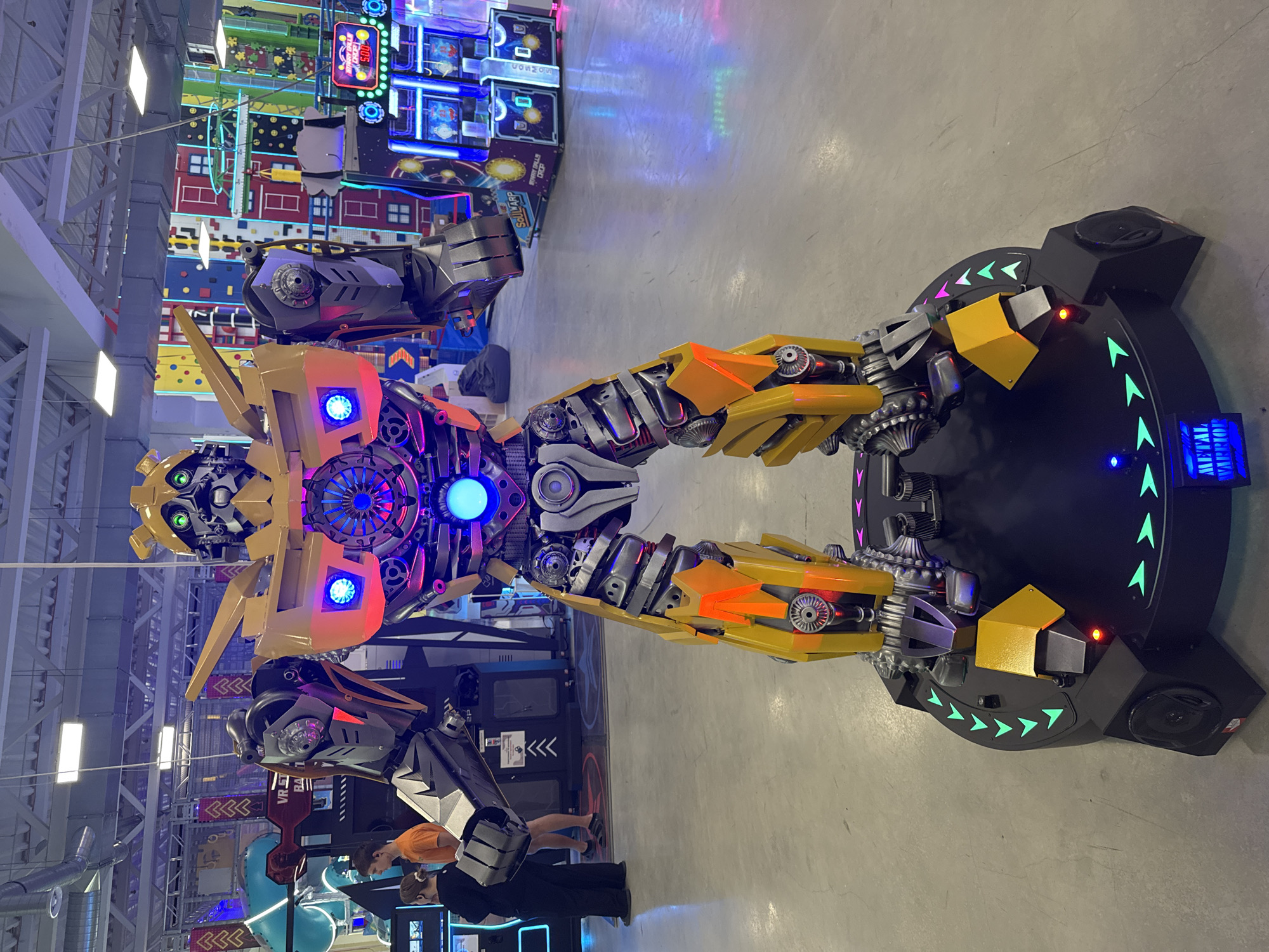 This image depicts a life-sized Bumblebee robot figure, likely an animatronic or costume, standing in an indoor entertainment venue. The robot is predominantly yellow and gray, with glowing blue and red lights integrated into its design, particularly on its chest and arms. It is positioned on a black circular platform with green arrow-shaped lights pointing inwards. In the background, various arcade games with colorful displays are visible, along with climbing walls adorned with city-themed murals. Two individuals, a woman and a man, are standing to the left of the robot, facing away from the camera. The overall environment suggests an arcade or amusement center. The location is identified as Orlivshchyna, Ukraine.