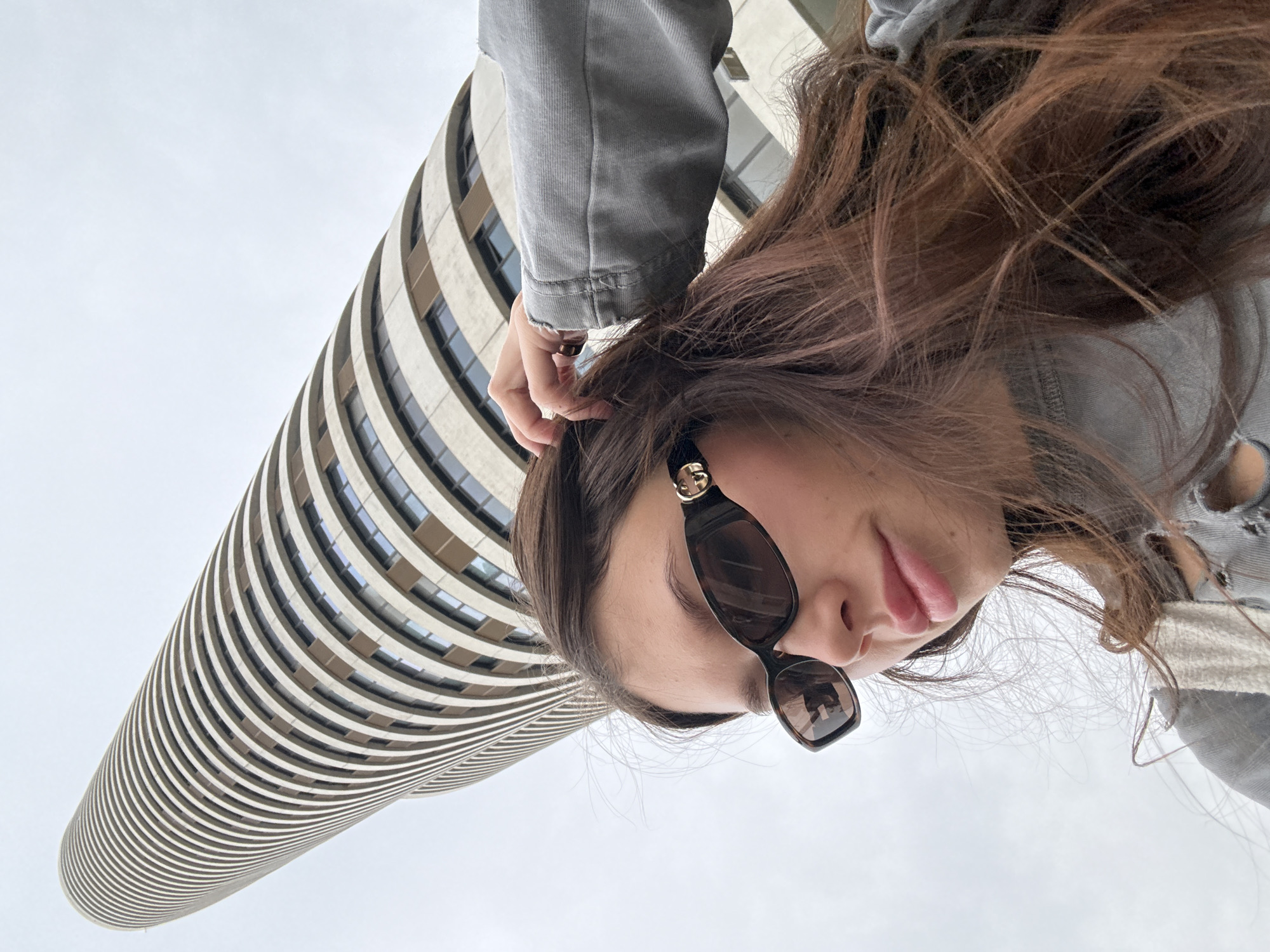A low-angle shot showcases a woman with brown hair and sunglasses standing beneath the Electra Tower in Tel Aviv, Israel. The tower's distinctive cylindrical shape with horizontal bands and evenly spaced windows is prominent against a pale sky. The woman is wearing a gray jacket and is holding her hair.