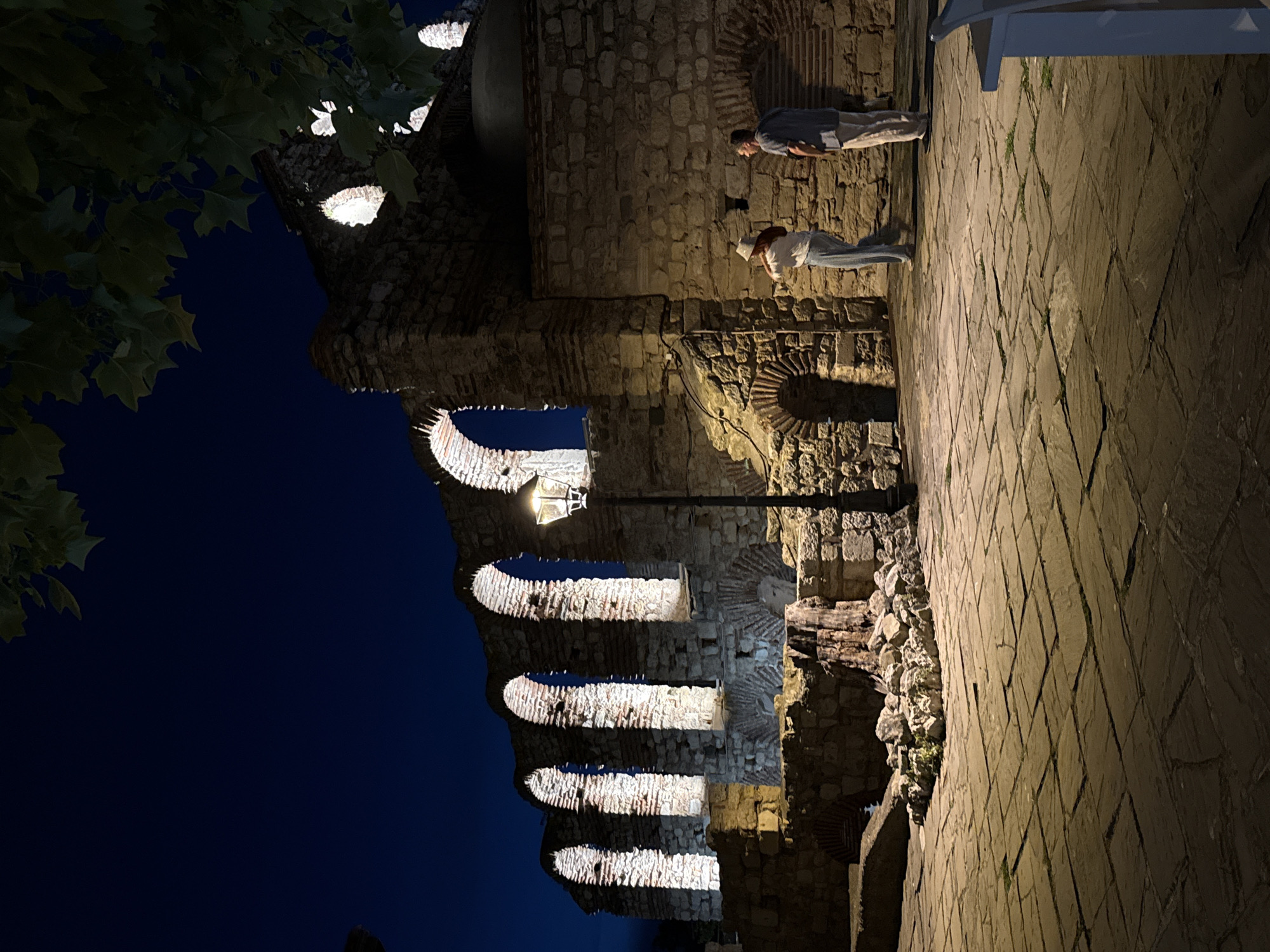 The image shows the ruins of the Church of St. Sophia in Nesebar, Bulgaria, at night. The stone structure features multiple arched openings illuminated from within. In the foreground, two individuals stand on a paved courtyard. A woman in a white hat and light-colored pants faces away from the camera, looking towards the ruins. A man in a light-colored shirt and pants stands beside her, also facing the ruins. A lamppost stands to the right of the ruins. The sky is dark blue, and tree branches with leaves are visible in the upper right corner.