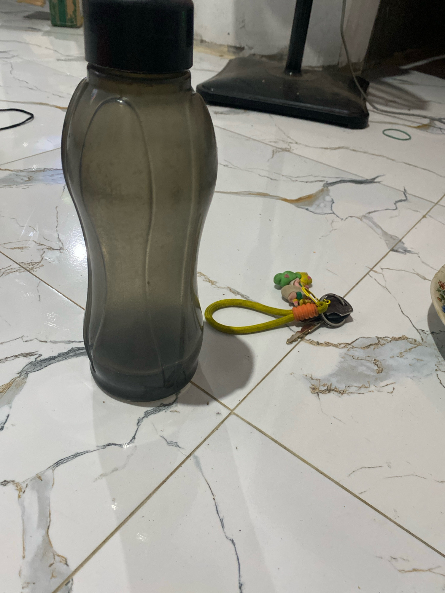 A darkly tinted, translucent plastic water bottle with a black cap stands upright on a white tiled floor with marble-like gray veining. To the right of the bottle, a yellow lanyard with a colorful cartoon charm and a key are attached. The background shows the base of a black floor fan and some electrical cords. The scene is indoors, likely in a residential setting in Planjan, Indonesia.