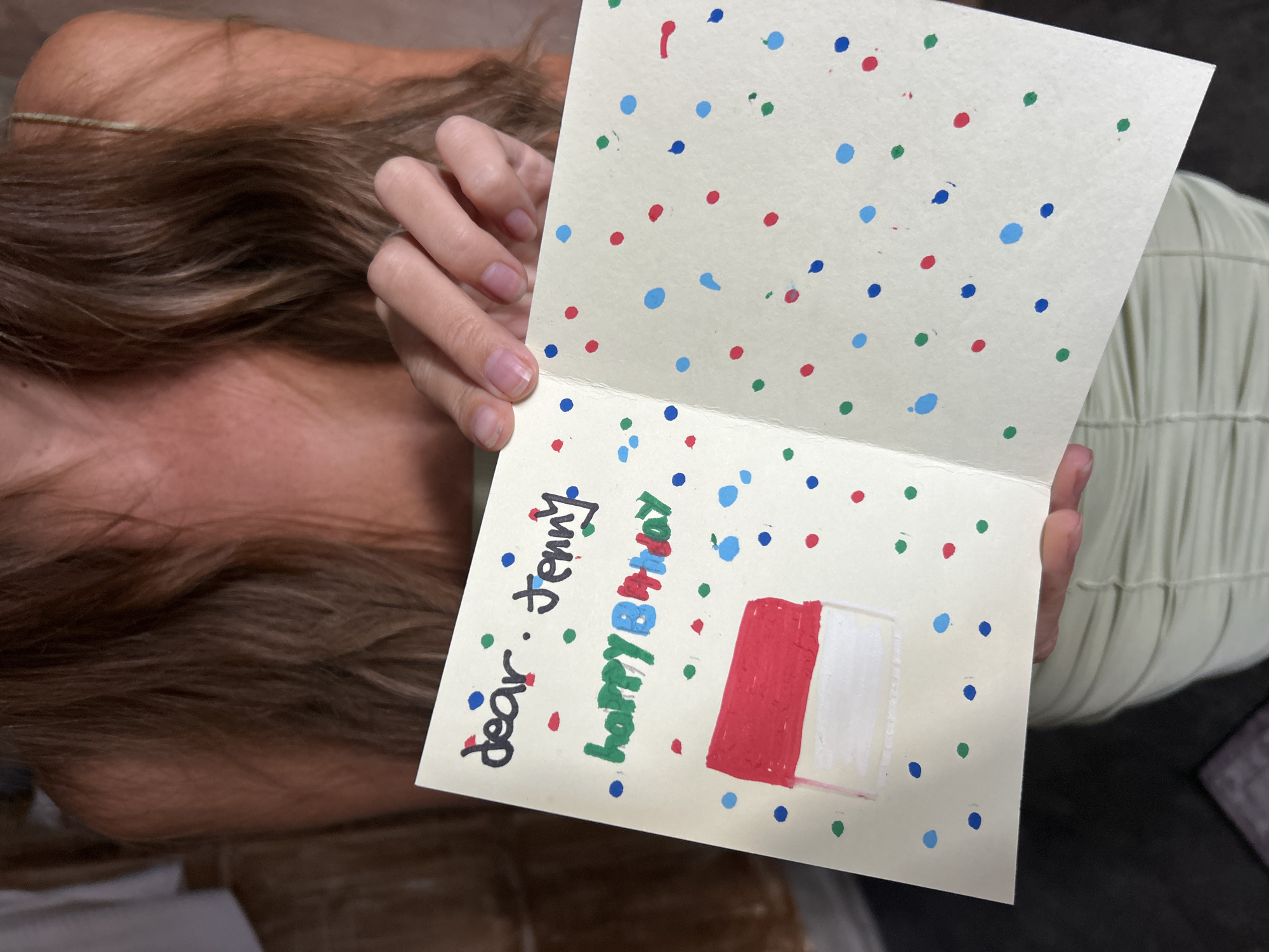A person is holding a homemade birthday card. The card is open and decorated with red, blue, and green polka dots. Inside the card, it reads "dear: Jenny happy B-Day". A drawing of a red and white rectangle, presumably a cake, is also visible. The card is held against a background that includes parts of the person's body, showing brown hair and a light green pleated dress. The location is North Kuta, Indonesia.