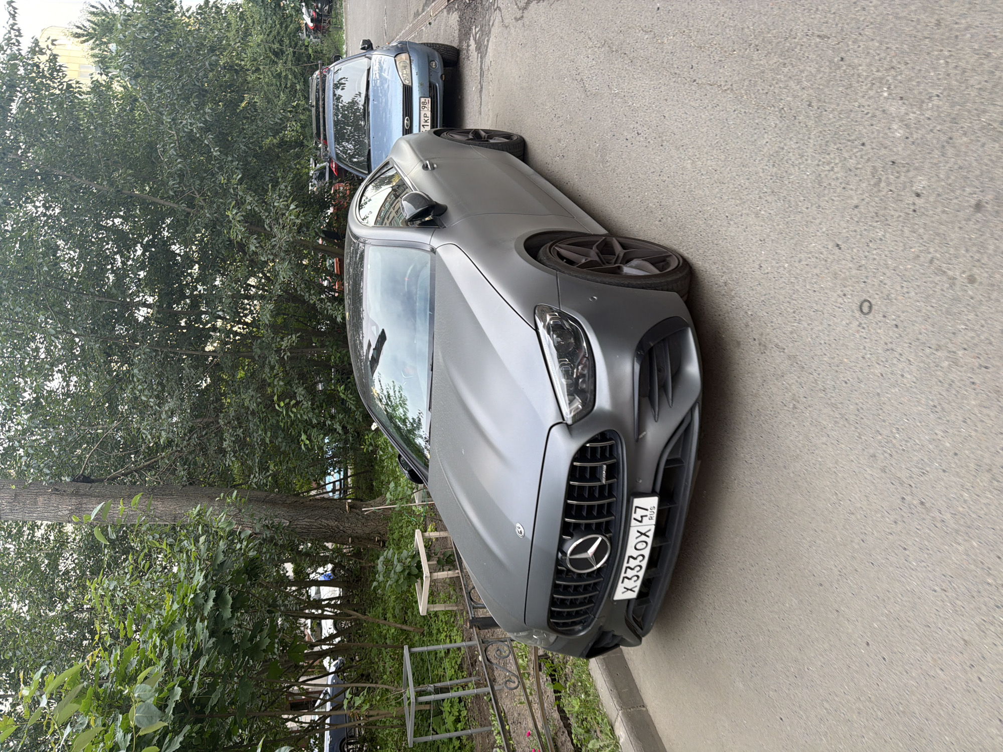 This media file shows two cars parked on a street next to a sidewalk and trees. The car in the foreground is a gray Mercedes-Benz with a black grille and silver wheels. The license plate is visible and reads "X333OX 47 RUS". The car in the background is a light blue sedan with a roof rack. The scene appears to be outdoors during the daytime, with dappled sunlight filtering through the trees. The overall setting is a residential or urban area with vegetation. There are no people visible in the image.