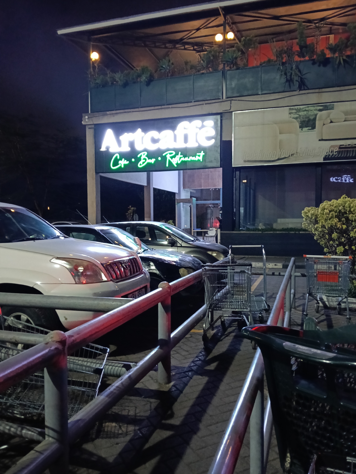 The image depicts the exterior of an Artcaffé establishment in Nairobi, Kenya, at night. The brightly lit "Artcaffé" sign is visible above the entrance, with "Cafe · Bar · Restaurant" written in green neon below. The restaurant has a second level with planters and lighting. To the right of the sign is a billboard advertising living room furniture and a smaller Artcaffé logo. Several cars are parked in front of the restaurant. A metal railing and a row of shopping carts are positioned in the foreground. The overall lighting suggests an urban, commercial area. The atmosphere is dark, with the main light sources being the restaurant's sign and other artificial lights.