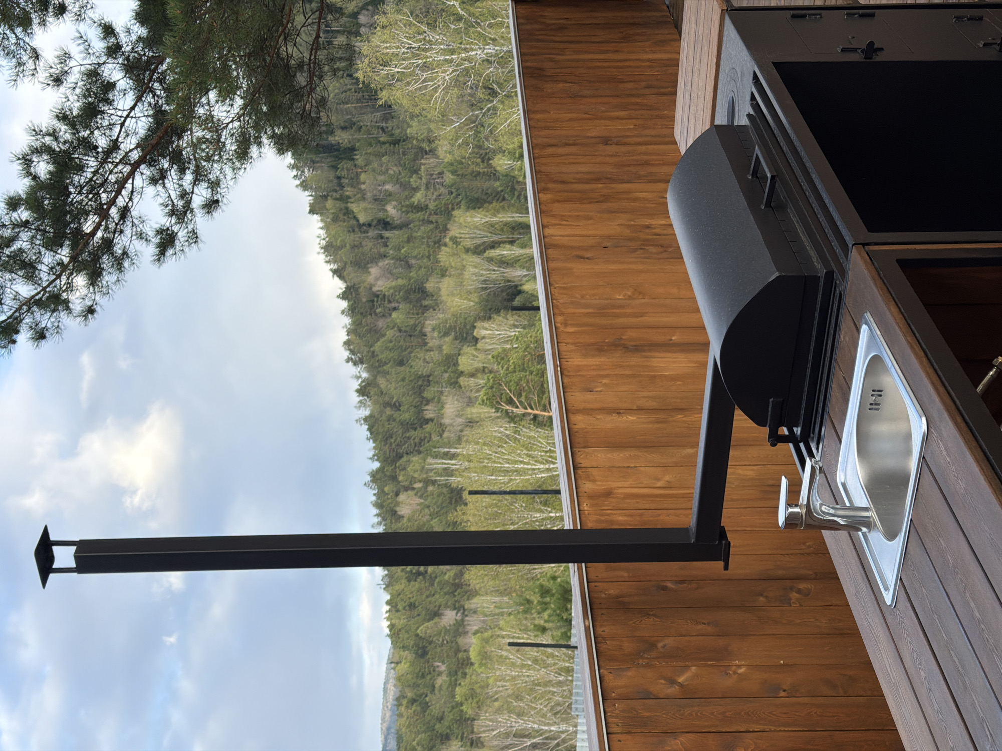 The image shows a metal outdoor grill and sink station on a wooden deck in a landscape. The grill is black with a cylindrical smoke exhaust and is positioned next to a stainless steel sink with a faucet. A black metal post with a flat cap extends from the grill towards the left, framing a view of a forest-covered hillside under a cloudy sky. The deck is made of stained wood planks. The environment includes a pine tree visible in the upper left corner and deciduous trees on the hillside. There are no people visible in the image. The location is likely in a natural setting near Манский, Russia, as indicated.