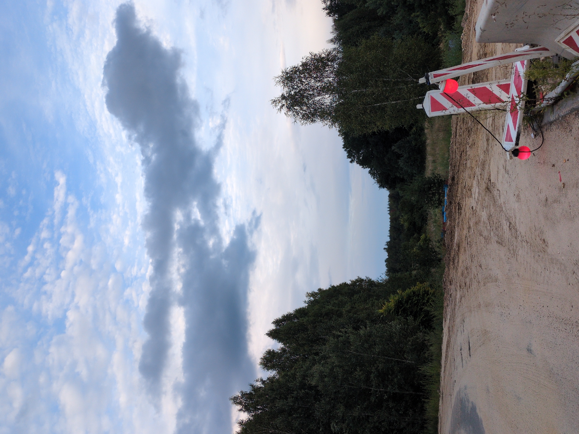 A dirt road curves to the left, bordered by a line of trees on the left and a construction site on the right. Two red and white striped barriers with red lights are positioned on the right side of the road. The sky above is filled with a mix of blue and grey clouds.