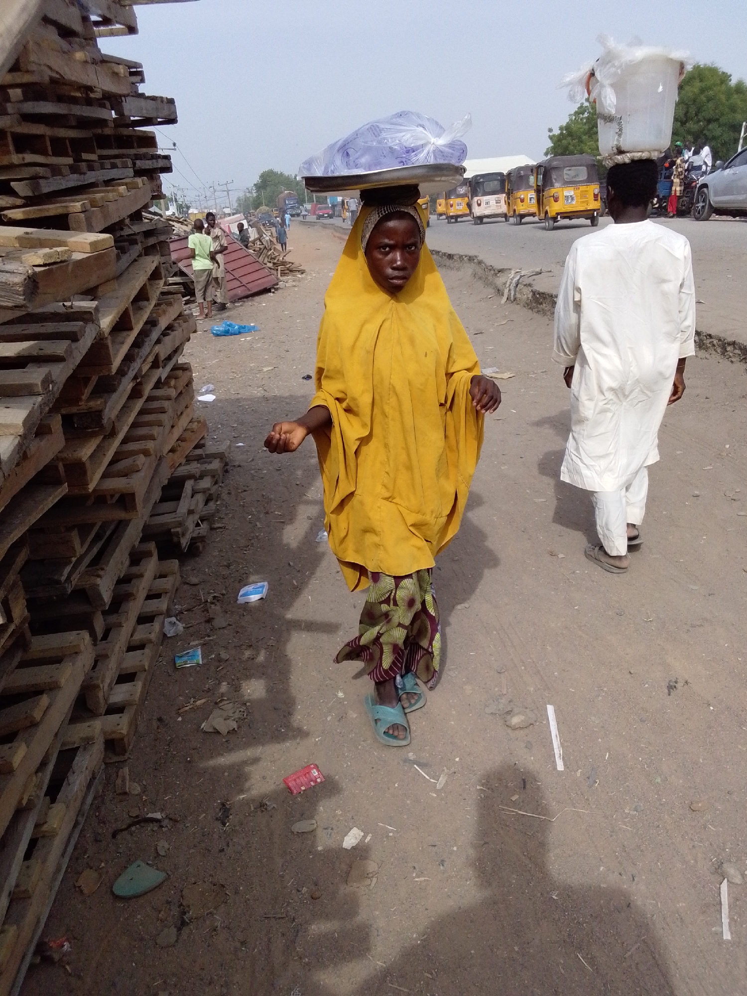 In Jimeta, Nigeria, a person wearing a yellow garment and patterned trousers walks toward the camera, carrying a tray covered with a blue plastic bag on their head. To the right, a second person in a white robe walks in the same direction, with a white bucket on their head. Behind them are several yellow auto rickshaws parked on the roadside. To the left of the first person is a stack of wooden pallets. Litter is scattered on the ground, which appears to be a dirt road. In the background, other people are visible along the road, and some buildings and trees can be seen in the distance. The sky is overcast.