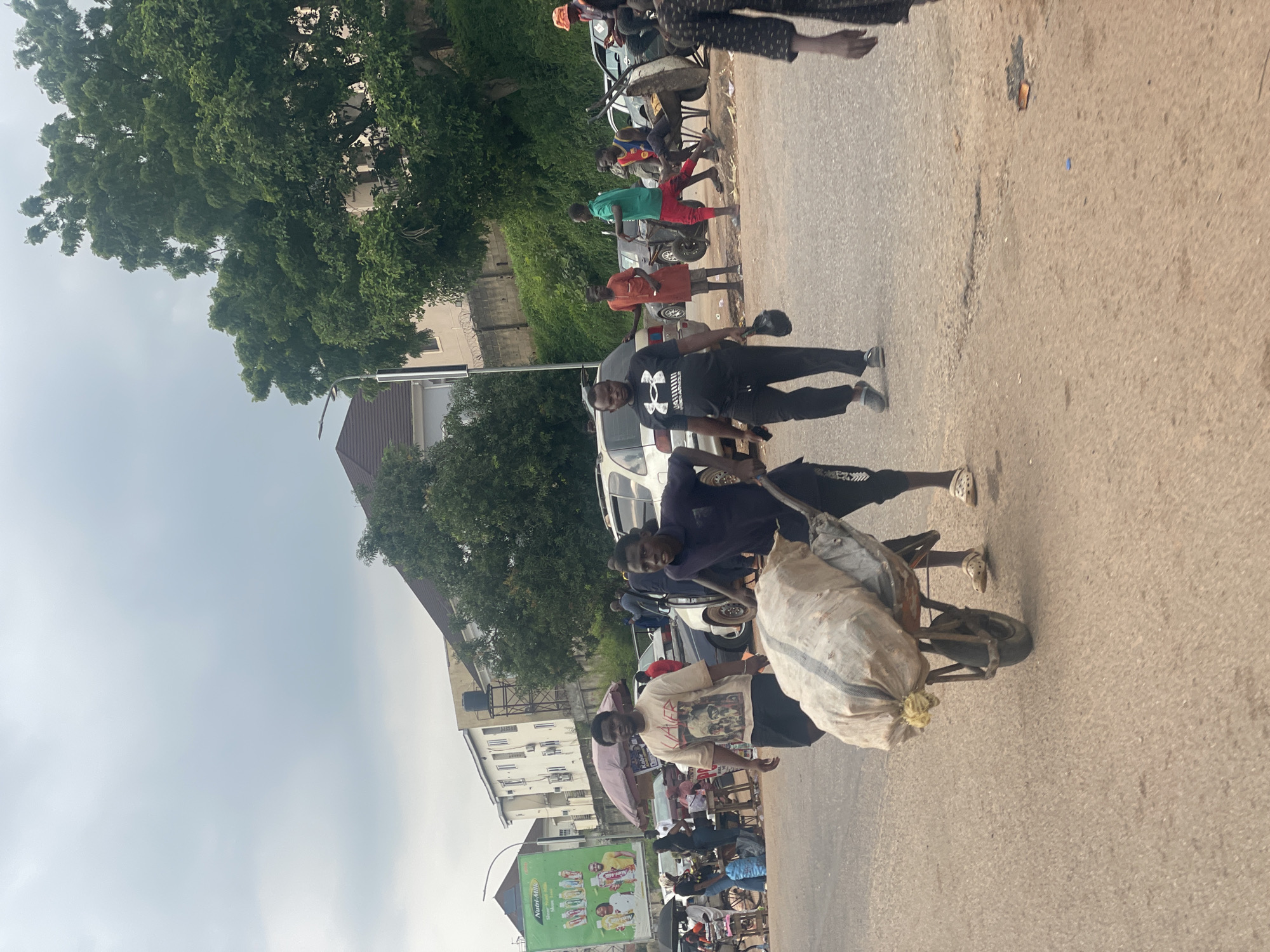 The image depicts a street scene in Abuja, Nigeria, on a cloudy day. In the foreground, a person is pushing a wheelbarrow loaded with a large, off-white sack. The wheelbarrow has one wheel and a metal frame. Behind the person with the wheelbarrow, another person is walking. Further in the background, several cars are parked, and a billboard for "Nutri-Milk" is visible on the left. A large, leafy tree dominates the right side of the frame. Several other individuals are scattered throughout the scene, some standing and others appearing to be walking. The environment appears to be an urban or semi-urban setting with paved roads and some buildings visible.