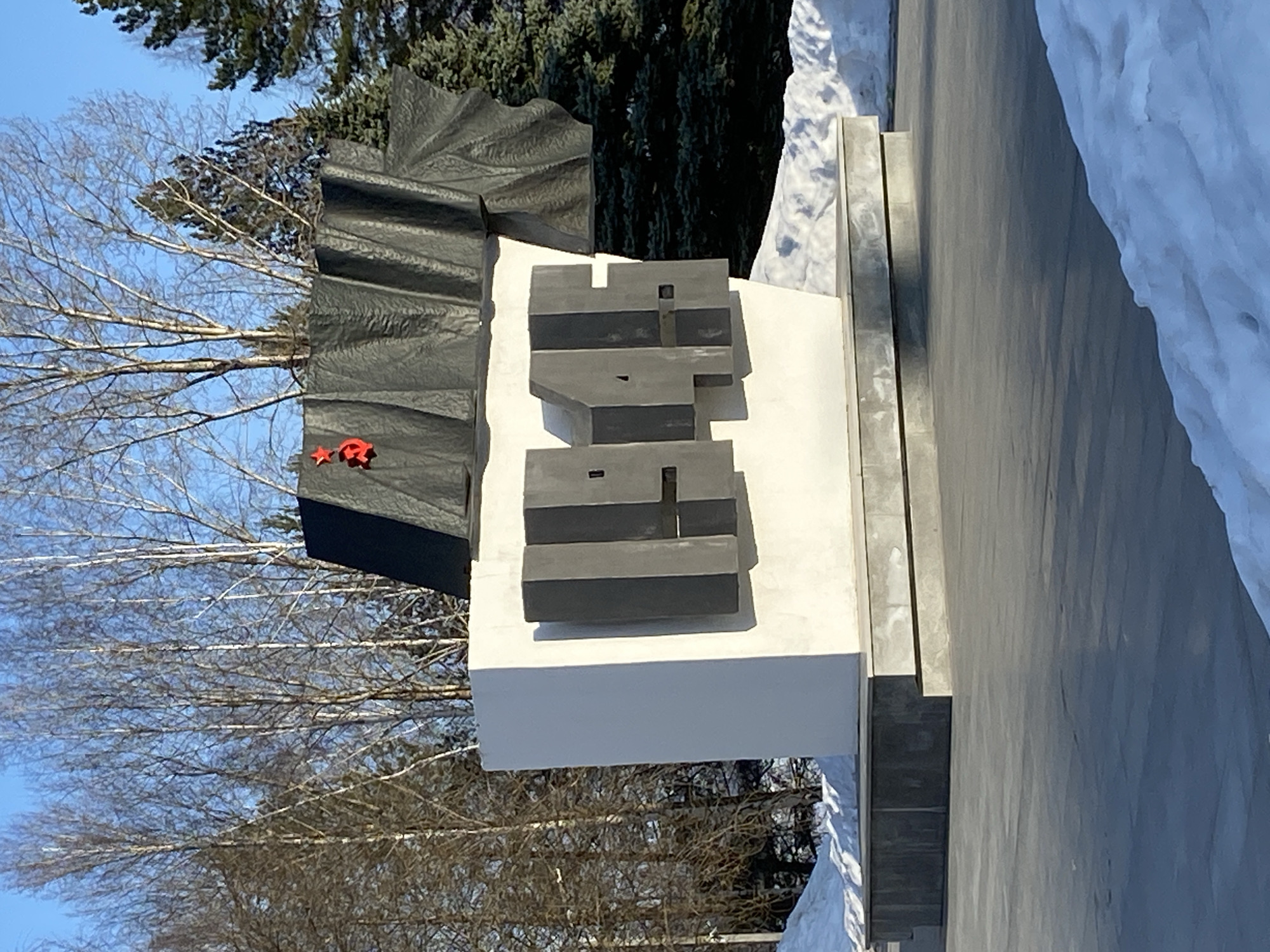 The image displays a monument situated outdoors in the city of Chaikovsky, Russia. The monument consists of a prominent white rectangular base, featuring the dark grey, raised numerical sequence "1941" and "1945" on its front face. Above this base, a dark, corrugated or stylized fabric-like structure is angled upwards, bearing a red star and a red hammer and sickle emblem on its upper left section. The monument rests on a multi-tiered grey concrete platform. The immediate foreground is a smooth, grey paved surface, with accumulations of snow visible along its edges and at the base of the monument. The background features a clear blue sky above a dense line of trees, including leafless deciduous trees with light bark and dark evergreen conifers. The scene is illuminated by direct sunlight, casting distinct shadows. No individuals or discernible actions are present.