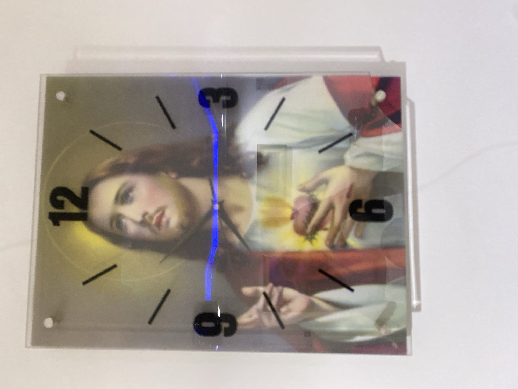 This is a photograph of a wall clock featuring an image of Jesus Christ. The clock has a clear acrylic face and black Arabic numerals marking the hours 12, 3, 6, and 9. The image of Jesus depicts him with a halo, looking upwards and holding his chest where his heart is visible and aflame. The clock is mounted on a white wall, and a blue light is reflecting off the acrylic surface near the 3 and 9 o'clock positions. The photograph does not provide any information about its location in Giginyu, Nigeria, or any other details of the environment beyond the clock and the wall.