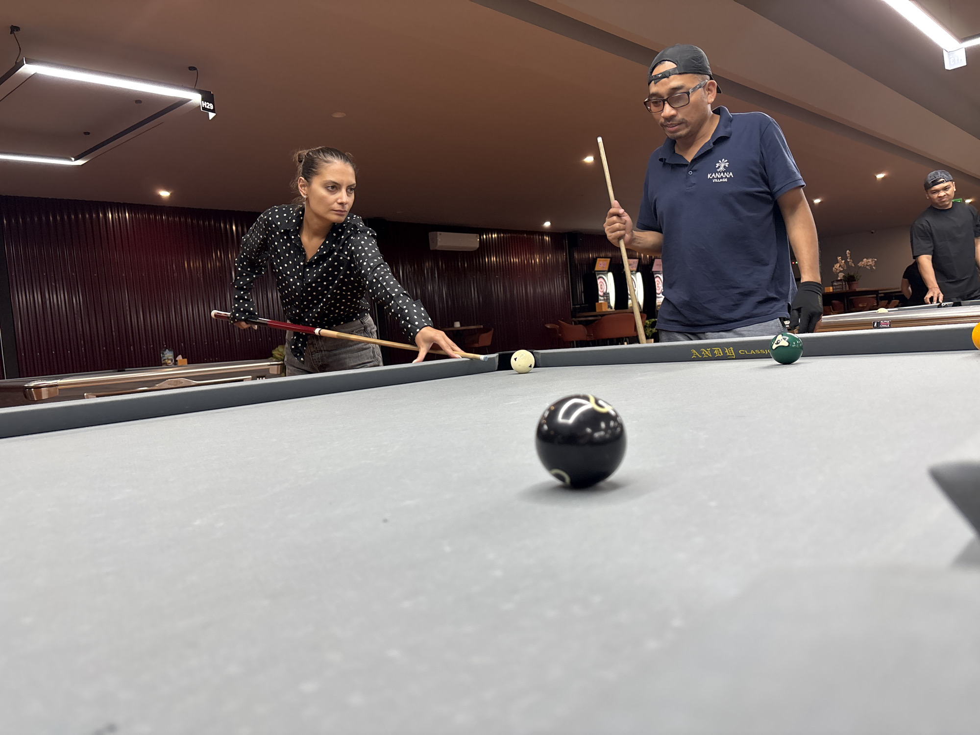 A woman in a black polka dot shirt and denim shorts is playing pool, aiming her cue at a white ball on a grey felt table. A man wearing a navy blue polo shirt, glasses, and a black baseball cap is standing to her right, also holding a cue. Another man in a black t-shirt and a black baseball cap stands in the background. The setting appears to be a dimly lit billiards hall, possibly in Kuta Selatan, Indonesia, with red wooden paneling on the walls and modern light fixtures on the ceiling. Other pool balls, including a black 8-ball, are visible on the table.