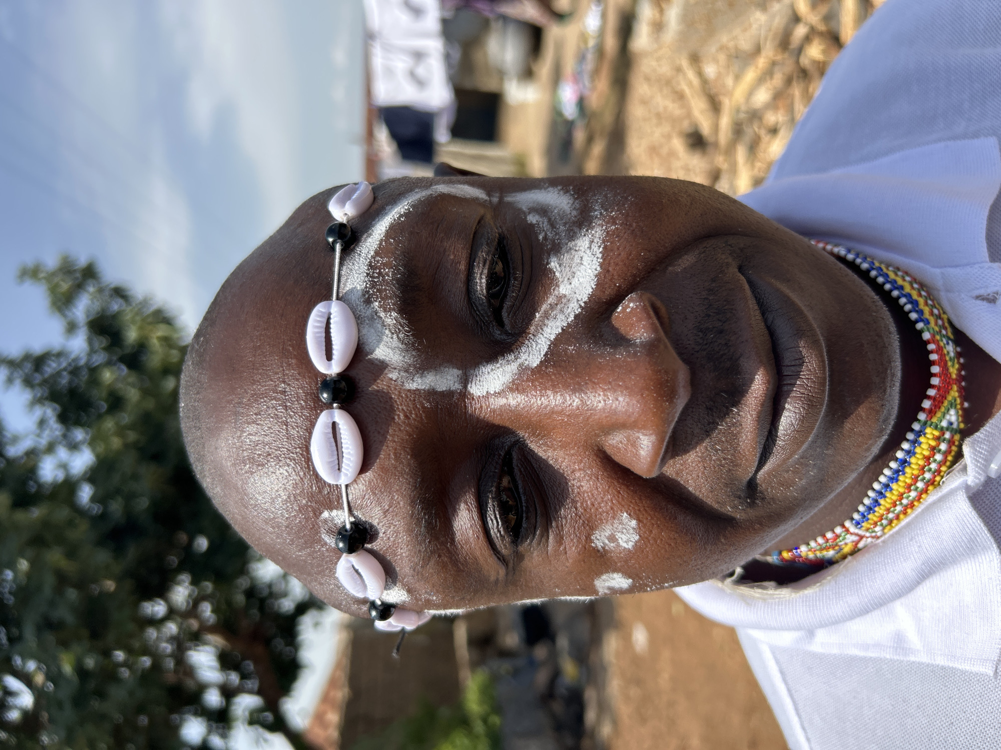This is a close-up, eye-level, portrait-style photograph of a dark-skinned man with a shaved head. He is adorned with white face paint, with a circular design on his left cheek and a wider, curved stroke across his forehead and extending down his temples. He wears a headband made of small white cowrie shells and black beads. Around his neck, he has a colorful beaded necklace. He is wearing a white polo shirt. The background is out of focus, showing a dirt ground, some dry stalks or corn husks, and structures with laundry hanging in the distance. The lighting suggests it is daytime, possibly sunny, with the sun casting shadows. The setting appears to be an outdoor, informal environment, likely within a residential area in Jos, Nigeria.