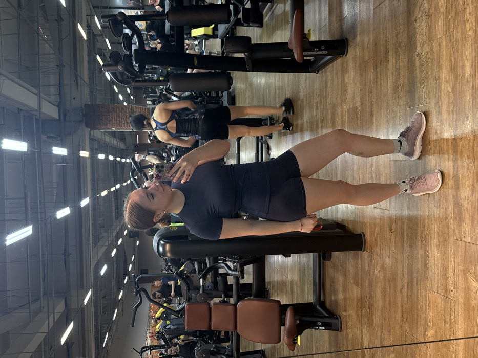 The image displays an indoor fitness center in Chaikovsky, Russia. The primary subject, a young adult female, stands in the foreground, angled slightly to her left, looking at her reflection in a large mirror. She holds a smartphone with a floral-patterned case in her right hand, positioned to take a photo. She is dressed in a dark blue short-sleeved athletic top and matching shorts, with her light brown hair pulled back. She wears white socks with black Nike swooshes and athletic shoes, one light pink on her left foot and one light grey on her right. A red string bracelet is visible on her left wrist, and a faint mark is present on her right shin.
In the middle ground, another female is seen from the back, wearing a blue patterned athletic top, black shorts, and a black waist trainer, with dark athletic shoes. Various other individuals are visible throughout the background, engaged with or near numerous black and metallic gym equipment, including weight machines with brown padded seats. The facility features light-colored wooden or laminate flooring, a high ceiling with exposed ductwork, and multiple rectangular fluorescent light fixtures providing illumination. A section of a brick wall is also visible in the background, reflected within the gym's extensive mirrors.