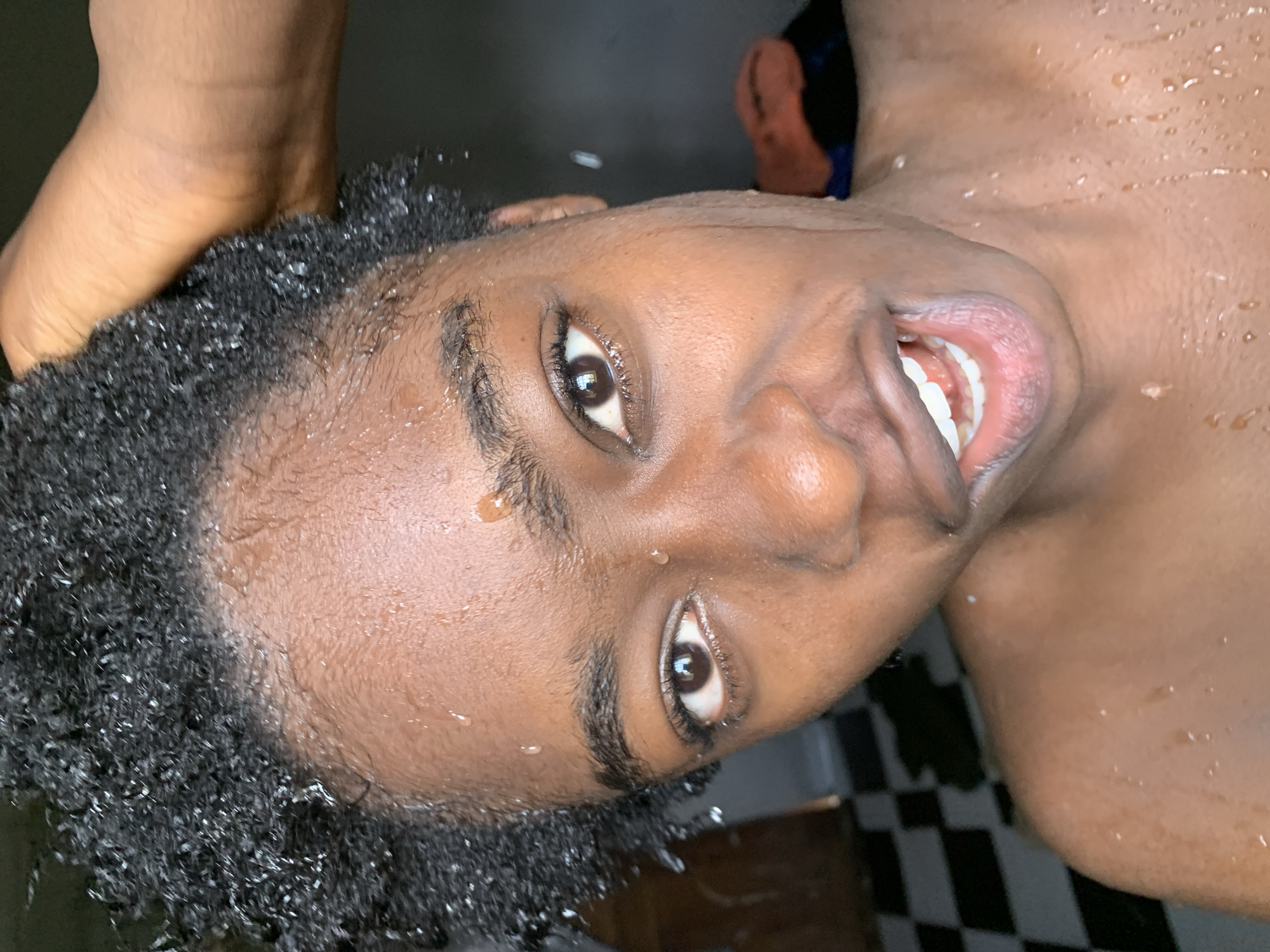 A close-up, waist-up shot captures a young person, appearing to be in a bathroom, with water droplets visible on their face, neck, and hair. The person has dark, curly hair, brown eyes, and is looking directly at the camera with their mouth open and teeth showing. Their head is tilted slightly, with a hand reaching up to touch their hair. The background is blurred, but a black and white checkered floor and a hint of a dark wall with an orange towel can be seen. The scene appears to be located in a residential setting in Rafin Par, Nigeria.