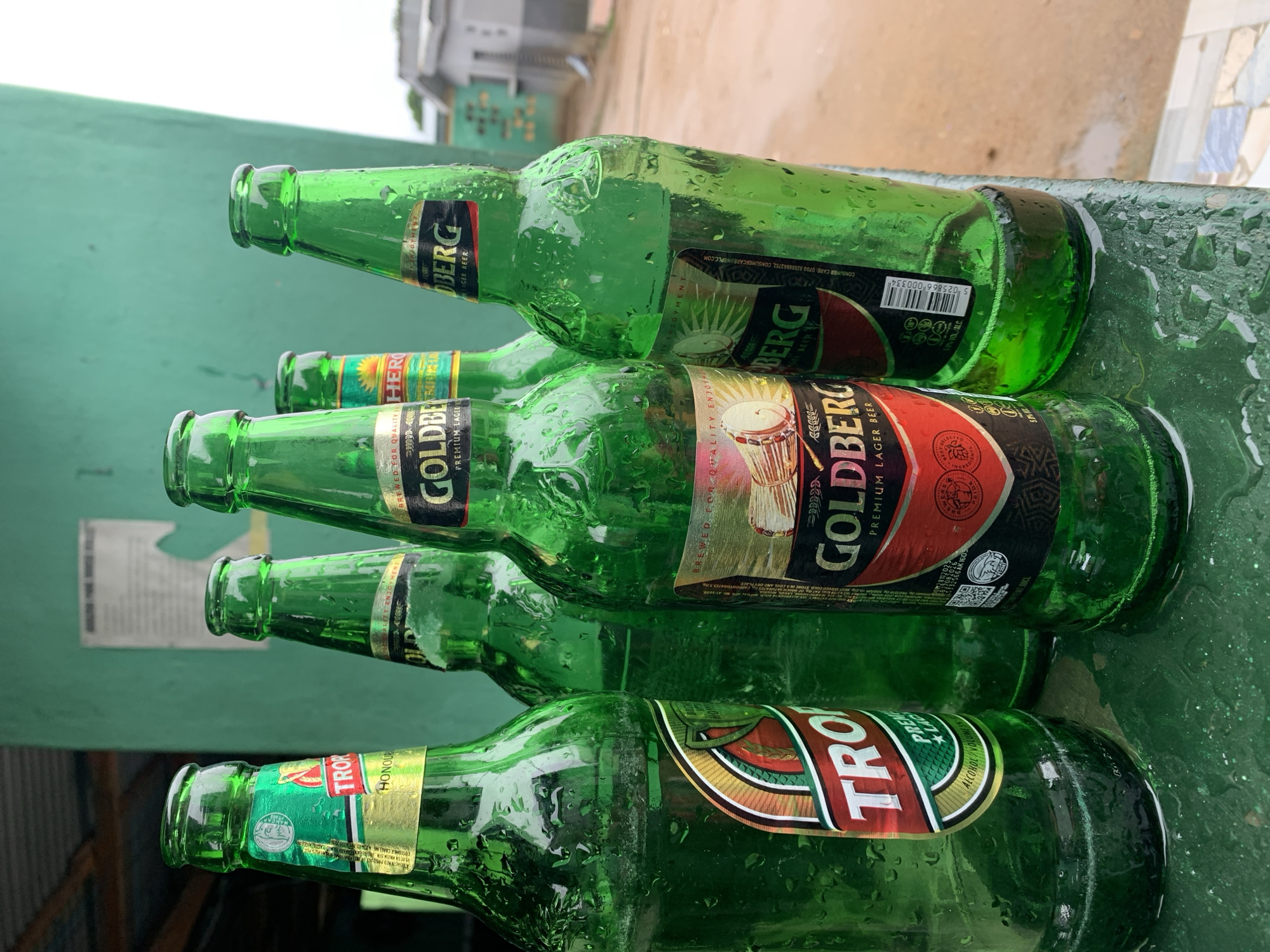 Five empty, wet green glass beer bottles lie horizontally on a wet, green surface in an outdoor setting at New Stadium, Nigeria. The bottles are covered in water droplets, suggesting recent rain or moisture. In the background, a blurred light-colored building and muddy ground are visible.
Three of the bottles are "Goldberg Premium Lager Beer," with prominent labels featuring a drum and wheat stalks. Visible text on these labels includes "GOLDBERG PREMIUM LAGER BEER," "BREWED FOR QUALITY ENJOYMENT," "CONSUMER CARE: 0700 823558662752, CONSUMERCARE INBPLC.COM," a barcode "025866 000334," and "VOL/ALC 5.5," with "CARBOHYDRATES 3.0" also partially visible.
One bottle is "Trophy Premium Lager," featuring a label with "TROPHY PREMIUM LAGER," and "HONOUR" along with "ALCOHOL VOLUME 5.5." Another bottle, partially visible, is a "Hero Lager Beer." The scene appears to be the aftermath of an event, with the empty bottles resting on what looks like a table or bench. The lighting indicates it is daytime.