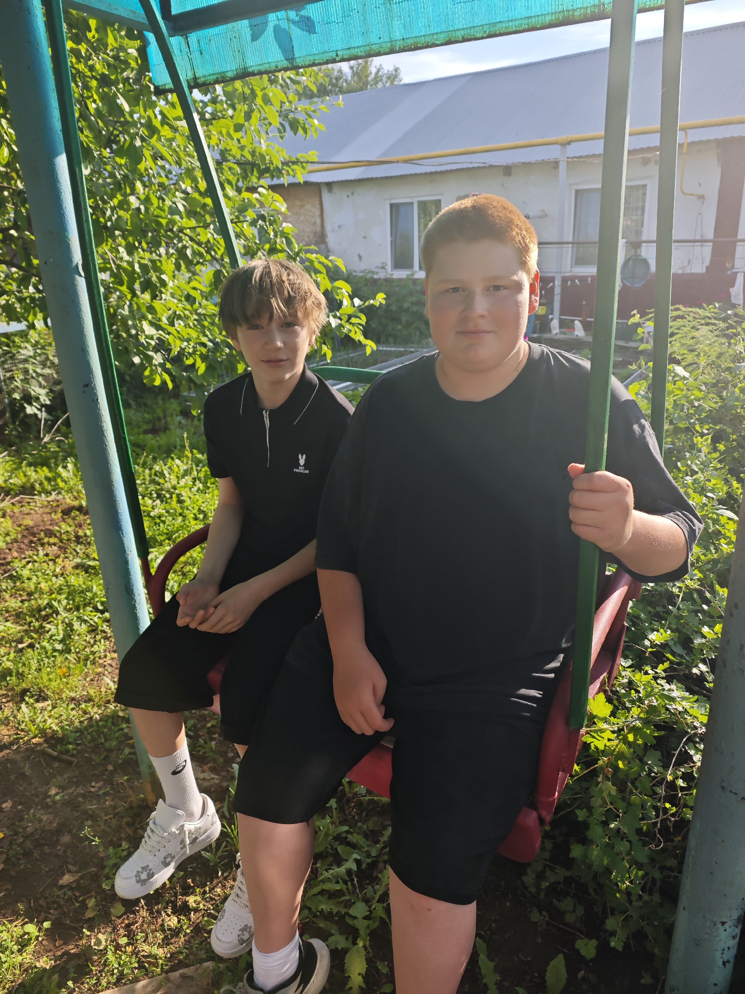Two young males, one with light brown hair and the other with reddish-blonde hair, are seated on a red swing. The male with light brown hair is on the left, wearing a black polo shirt and black shorts, with white sneakers. The male with reddish-blonde hair is on the right, wearing a black t-shirt and black shorts, with white sneakers. They are under a turquoise canopy, which is supported by green metal poles. Behind them, a light-colored building with a grey roof is visible. The area around the swing is overgrown with green vegetation and dry dirt. The sun is shining, casting light and shadows. This scene appears to be in the backyard of a residential property. Based on the provided context of the city of Фоки, Russia, this is likely a rural or suburban residential setting within that location.