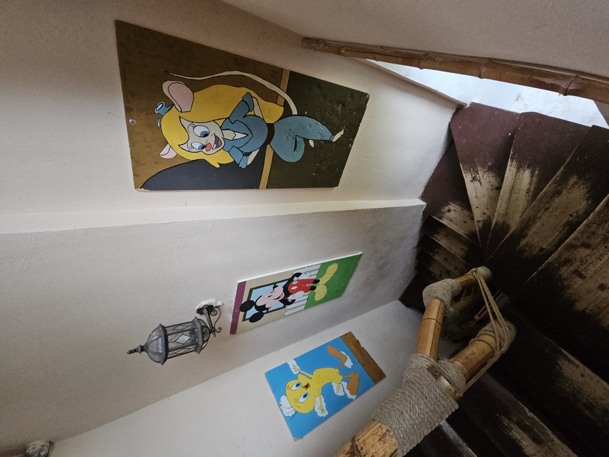 The image depicts a spiral staircase in a residential building, likely in Ulitkino, Russia. The staircase is constructed of dark wood with a maroon-colored coating on the steps. A bamboo railing, wrapped with thick rope, lines the right side of the stairs. Three cartoon-themed paintings are mounted on the off-white wall. The painting on the far left features the character Tweety Bird on a bright blue background. To its right, a painting depicts Mickey Mouse standing behind a white picket fence. The largest painting, on the far right, shows a female mouse character, likely Gadget Hackwrench from Chip 'n Dale Rescue Rangers, wearing a blue jumpsuit. An old-fashioned lantern is mounted on the wall above the Mickey Mouse painting. The lighting suggests it may be daytime, with natural light filtering from an unseen source. The overall impression is a quirky and personalized decorative style for a stairwell.