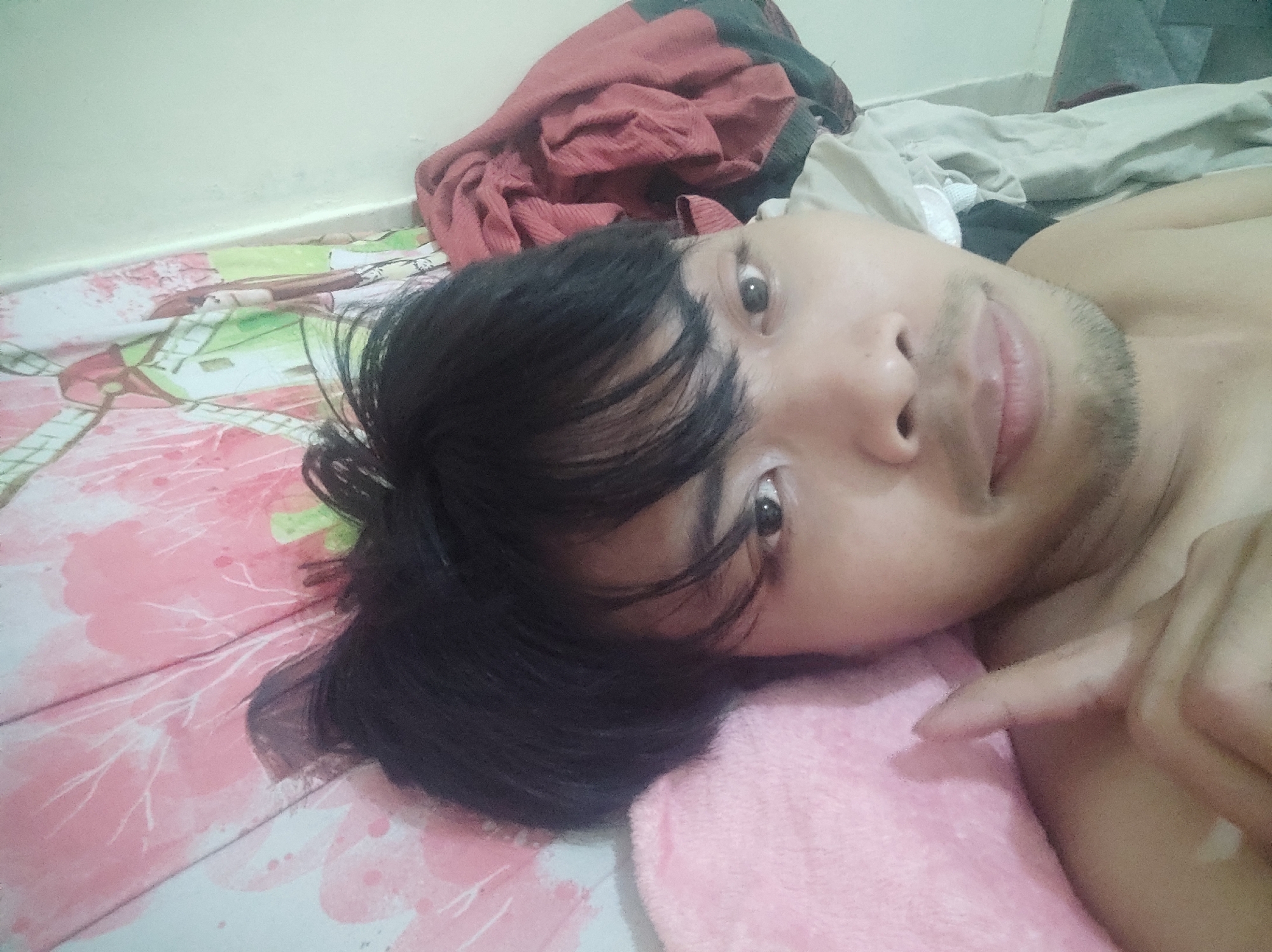 The image captures a close-up, selfie-style shot of a person lying on a bed. The person has dark hair that falls across their face, obscuring some of their features. Their eyes are visible and looking towards the camera. The person appears to be lying on a pink and white patterned bedspread, with a pink blanket or pillow positioned near their head. In the background, there are crumpled pieces of fabric, appearing to be clothing or blankets, in shades of red and dark grey, against a pale green wall. The scene suggests a domestic setting, potentially a bedroom. The presence of a bed, bedding, and personal clothing items indicates an indoor environment. The lighting is soft and diffused, typical of indoor lighting conditions. The image is taken from a low angle, consistent with a selfie taken while lying down. The location within the city of Trihanggo, Indonesia, is not directly discernible from the visual elements of the image itself.