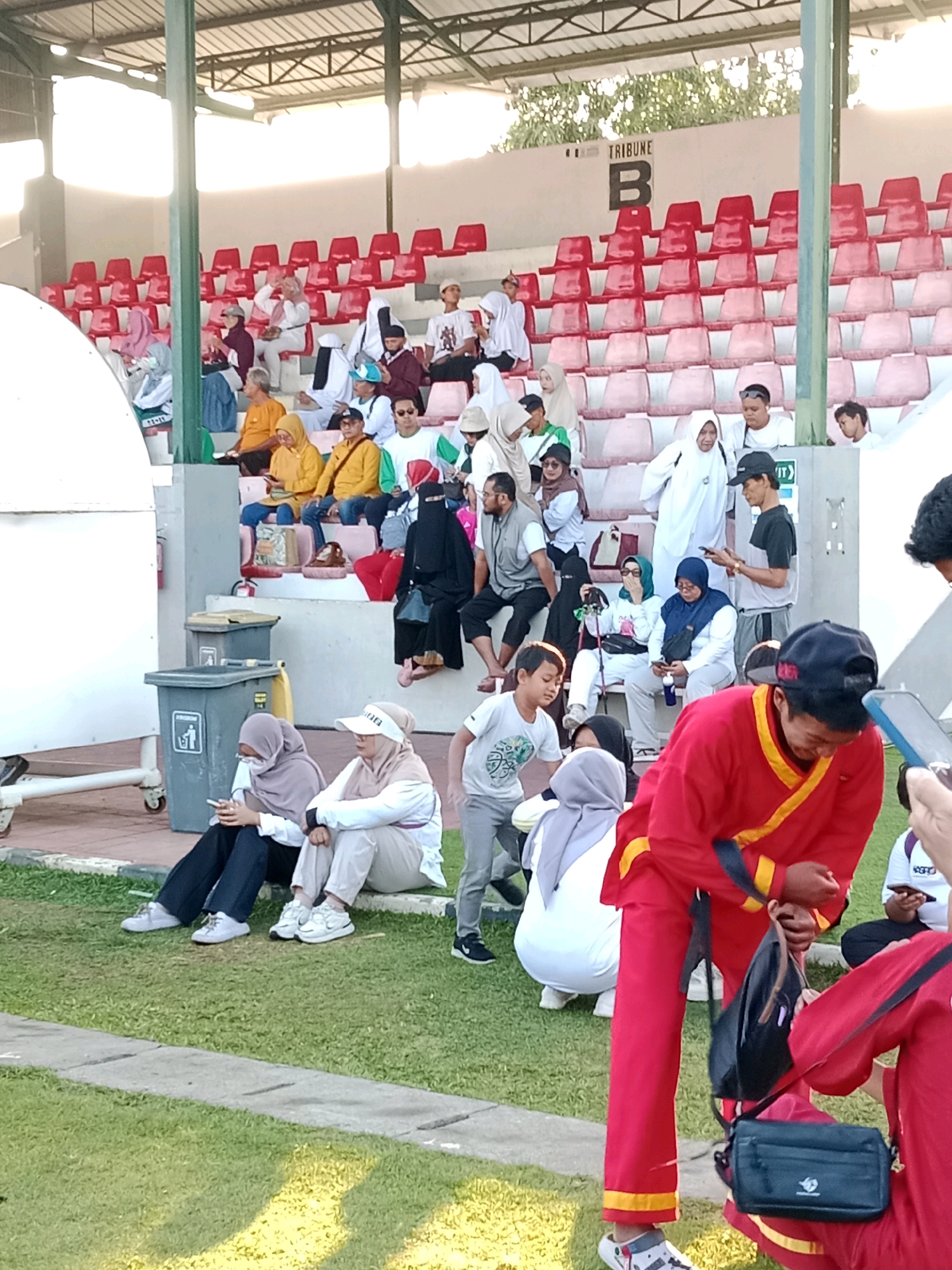 The image depicts a crowd of people, many wearing headscarves, gathered in the stands of a sports stadium in Surakarta, Indonesia. The stadium features rows of red and light-colored plastic seats, with a "TRIBUNE B" sign visible in the background. Some individuals are seated in the stands, while others are on the grass in front of them. A young boy in a white t-shirt with a green graphic is visible in the foreground, interacting with someone partially out of frame. Two trash bins are situated near the stands. The overall lighting suggests it is daytime, possibly late afternoon, with shadows cast by the stadium structure. The people are dressed in casual attire, with some wearing traditional Indonesian clothing. The scene appears to be a public gathering, possibly a sporting event or a cultural festival.