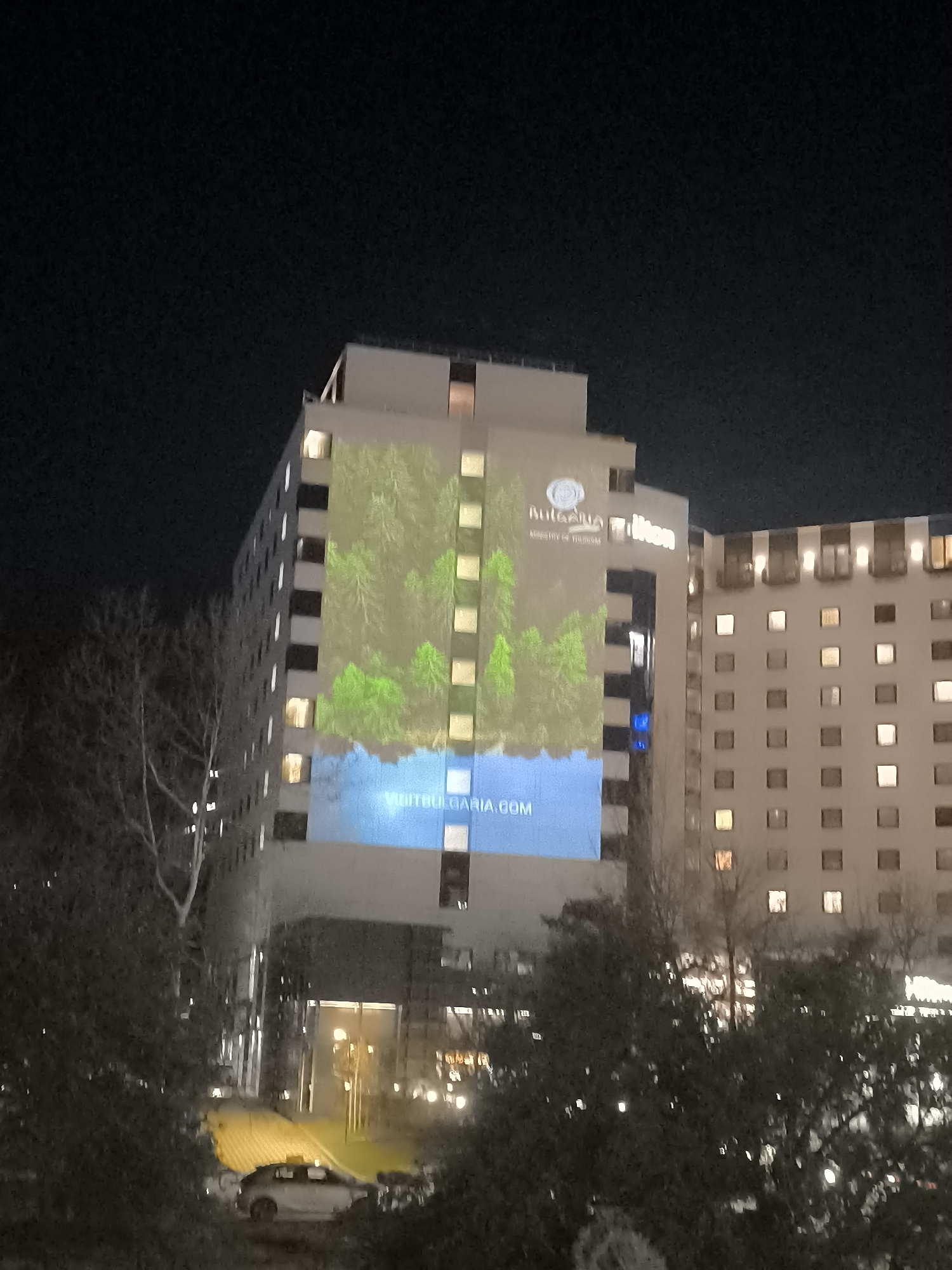 The image shows a view of a building in Sofia, Bulgaria, at night. On the side of the building is a projection featuring the text "Bulgaria Ministry of Tourism" at the top, an image of a forest, and the website "VISITBULGARIA.COM" at the bottom. The building is the Hilton Sofia, as indicated by the Hilton logo. To the left of the Hilton is another building. In the foreground are trees and what appears to be a car parked on the street.