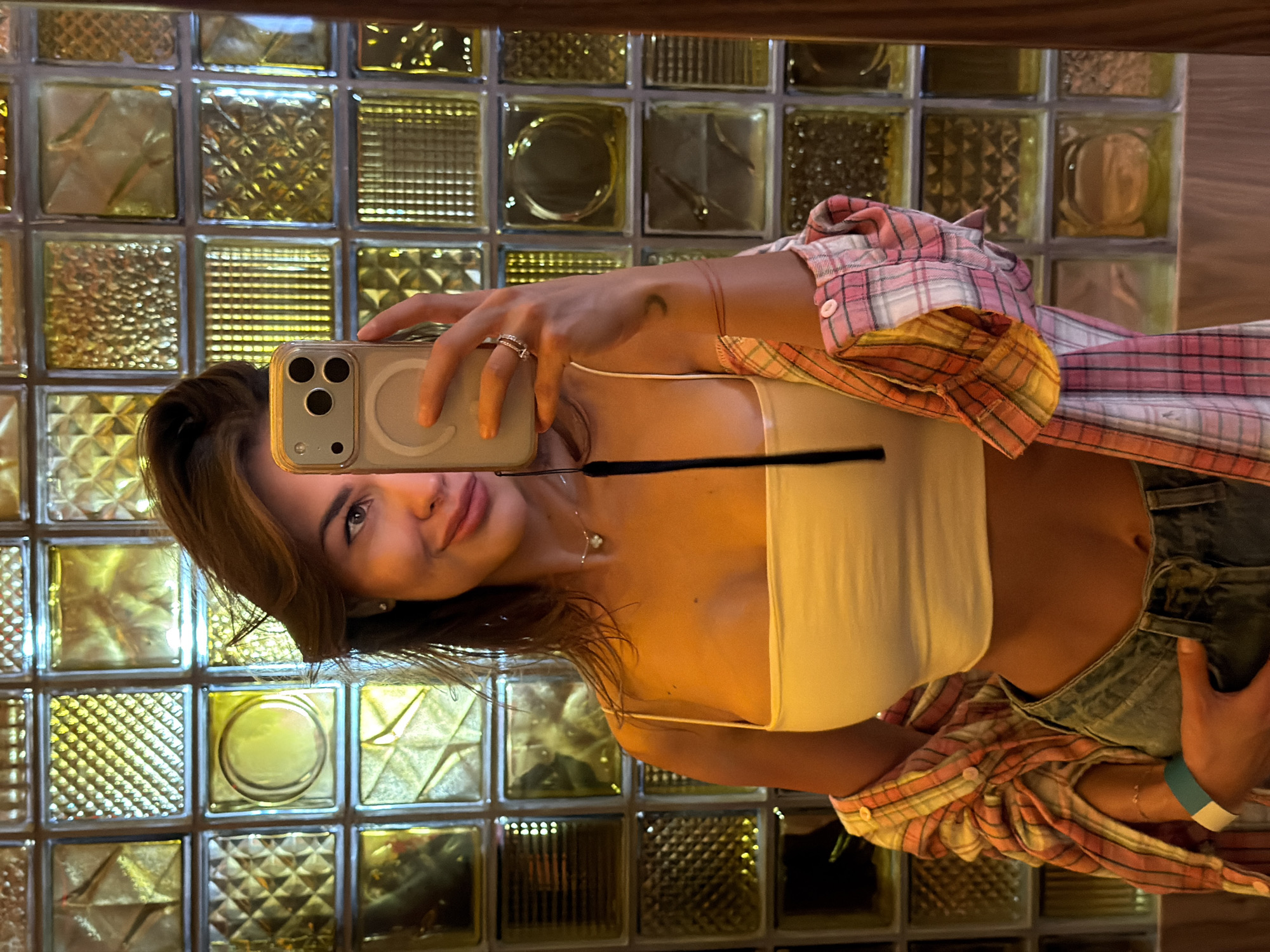 A woman is taking a selfie in front of a textured glass block wall. She is holding a smartphone in her right hand. She is wearing a white crop top, a pink and orange plaid shirt, and blue jeans. She is also wearing jewelry, including rings and a necklace. The background is a wall made of glass blocks with a textured pattern. The lighting appears to be soft and warm. The location cannot be identified as being in Krasnoyarsk, Russia based on the visual information.