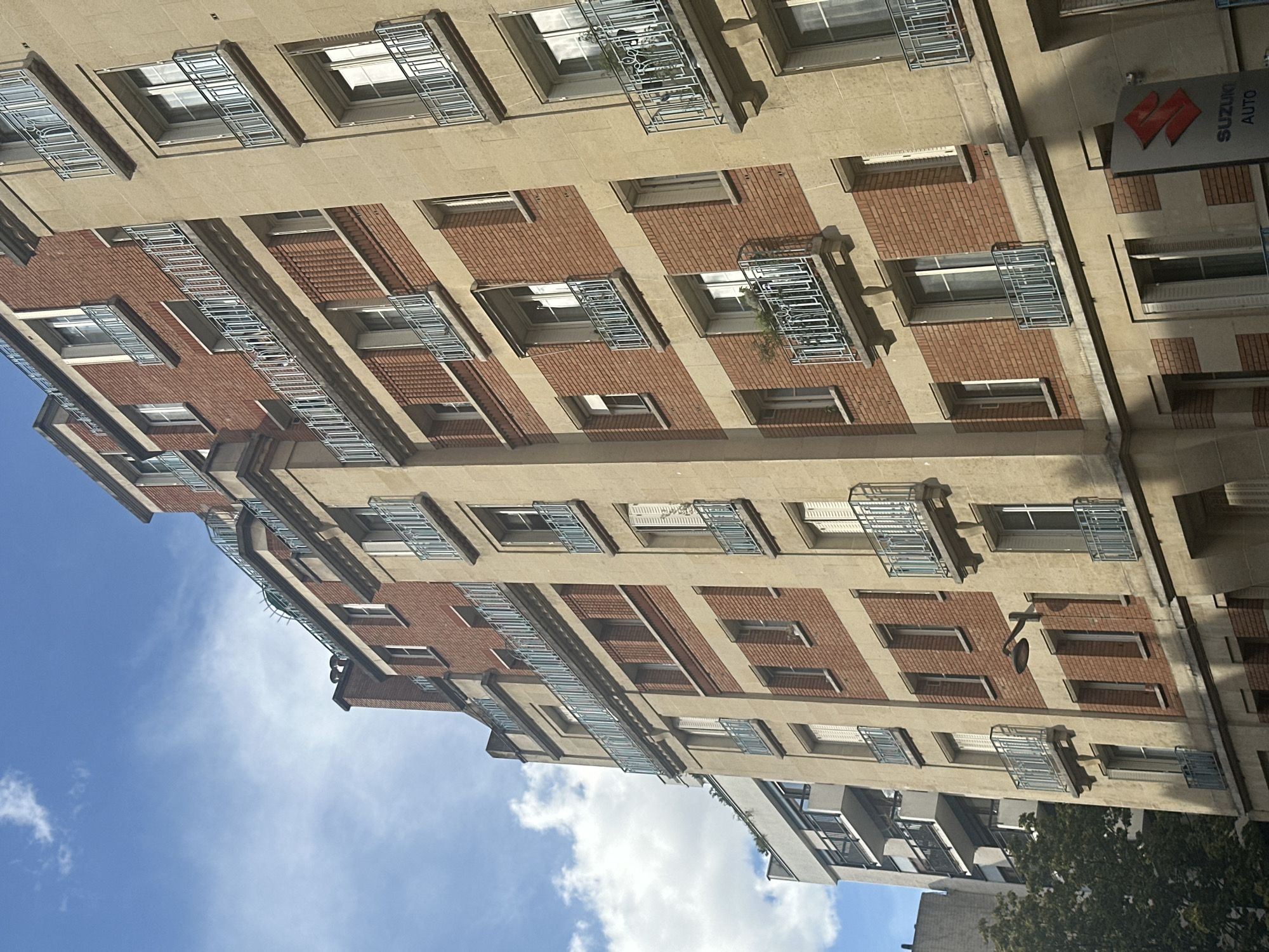 The image shows a section of an apartment building in Paris, France, under a partly cloudy blue sky. The building is constructed with red brick and cream-colored stone accents. It features multiple stories with regularly spaced windows, many equipped with light blue decorative balconies. Some balconies have small plants. A black street lamp is mounted on the building's facade. On the lower right, a Suzuki Auto sign is visible. The perspective is angled upwards, showcasing the height of the building and its architectural details.