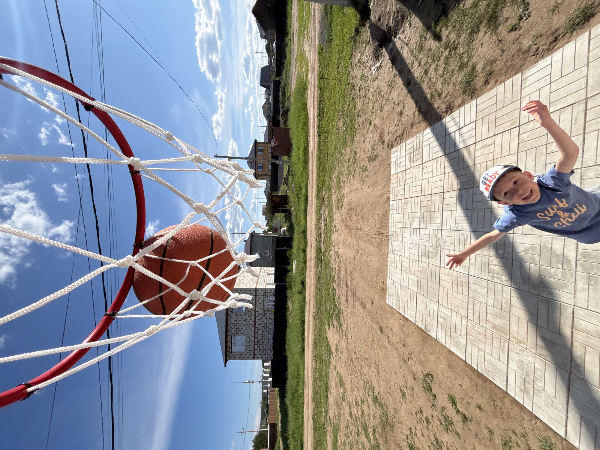 The image, taken in Chaikovsky, Russia, depicts a celebratory moment on a sunny day. A basketball is captured mid-air, having just passed through a red basketball hoop fitted with a white net, viewed from a low-angle perspective looking upwards. Directly beneath the hoop, a young boy, estimated to be between 5 and 8 years old, stands on a light-colored, patterned paved area, his arms outstretched in apparent excitement or triumph, and a wide smile directed towards the camera. He is wearing a blue short-sleeved t-shirt with "Surf & chill" printed on it and a white baseball cap with "GET READY" visible on the front.
The paved area, composed of rectangular tiles arranged in a herringbone pattern, serves as a partial court. It is surrounded by undeveloped ground, consisting of dirt and sparse patches of green grass. In the background, under a clear blue sky dotted with scattered white cumulus clouds, several residential structures are visible. These include at least two-story houses, one prominently built with light-colored concrete blocks and dark roofing. Overhead, multiple black power lines traverse the sky, connecting unseen utility poles. The scene is brightly lit by direct sunlight, casting distinct shadows on the paved surface and from the hoop.