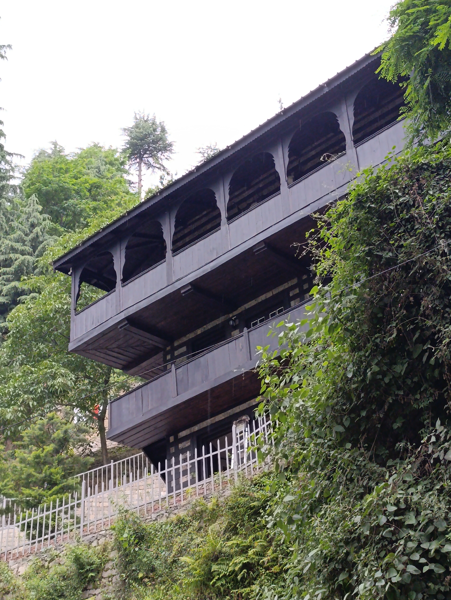 This image showcases a traditional multi-story wooden building situated on a steep, verdant hillside. The architecture features prominent dark brown wooden verandas or balconies on at least two levels, with the upper balcony distinguished by decorative arched openings. The building's design appears well-integrated into the natural slope.
Lush green vegetation heavily surrounds the structure. Dense trees, including conifers and broadleaf species, form a backdrop and flank the building. Thick vines and bushes cascade down the hillside in the foreground and along the right side of the image, some appearing to grow directly on the stone-faced terrain. A white picket fence runs along a lower section of the property, above a stone retaining wall.
The scene depicts a tranquil, natural setting, likely in a mountainous or hilly region. The sky is overcast, casting a soft, diffused light over the landscape, which could indicate a cloudy day, early morning, or late afternoon. No people or specific activities are visible, emphasizing the serene and architectural beauty amidst nature. The traditional wooden construction and mountainous environment are characteristic of the provided location context of Naggar, India. There is no visible text in the image.